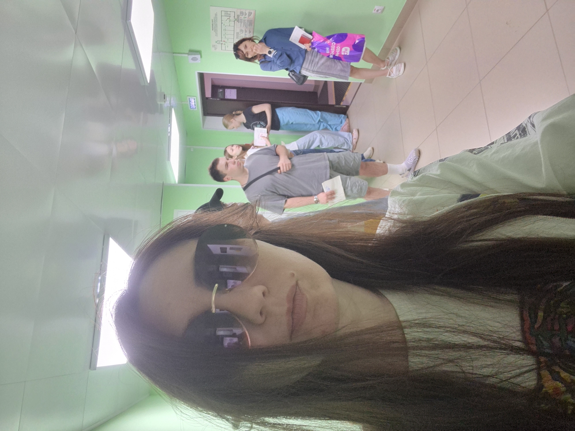 The image presents a selfie of a woman wearing sunglasses, with her hair obscuring part of the frame. In the background, a line of four individuals can be seen standing in a hallway. The first individual, a man, is dressed in a grey shirt and shorts, holding a document. Next to him, a woman in a white shirt also holds a document. Following them is another woman wearing a black shirt and blue pants. At the end of the line is a woman in a blue jacket and grey skirt, holding a pink bag. The hallway has green walls, and a directional sign in Cyrillic is visible above one of the doorways. The location is the city of Unknown, Unknown.