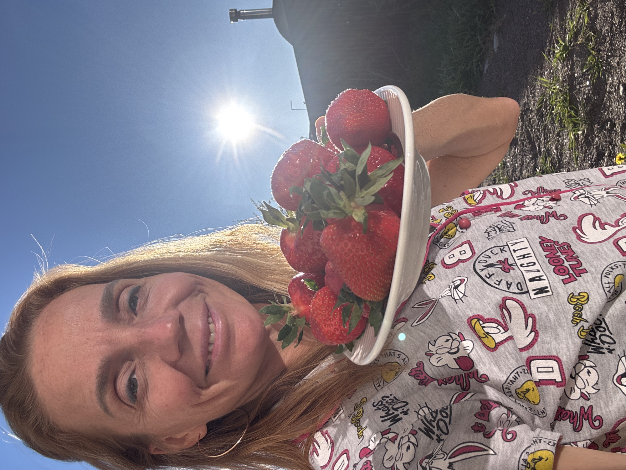A person is holding a bowl of strawberries in an outdoor setting. The individual, presumably a woman, is smiling and wearing a light gray pajama set patterned with cartoon characters. She has long blonde hair. The strawberries are bright red with green stems, filling the bowl. The background includes a bright blue sky with the sun shining, casting a lens flare effect. Parts of a dark object that could be a structure or equipment are visible in the upper left. The ground appears to be a mix of grass and soil. The location is unspecified as the image lacks indicators to pinpoint it to Steklyannyi, Russia.