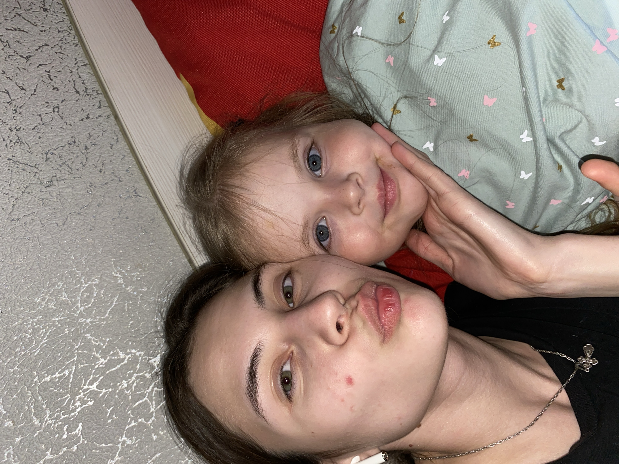 This close-up selfie depicts two females, an adult and a young child, lying side-by-side, likely in a casual indoor setting such as a bed or couch, in Votkinsk, Russia.
The adult, in the foreground, has dark hair and green eyes, posing with puckered lips in a "duck face" expression. She wears a dark top, a silver chain necklace with a small cross pendant, and a white earbud in her left ear. A small red blemish is visible on her right cheek.
The child, positioned behind the adult, has light brown hair and blue eyes, and looks towards the camera with a gentle, slightly smiling expression. The adult's right hand softly cups the child's face. The child wears a light green top patterned with pink, white, and gold butterflies.
The background includes a red fabric, possibly a pillow or blanket, behind both subjects' heads, and a white, vertically textured surface adjacent to it. A light grey wall with a distinct, abstract crack-like texture is visible behind the adult's head. The scene suggests a relaxed, affectionate moment. No text is visible in the image.