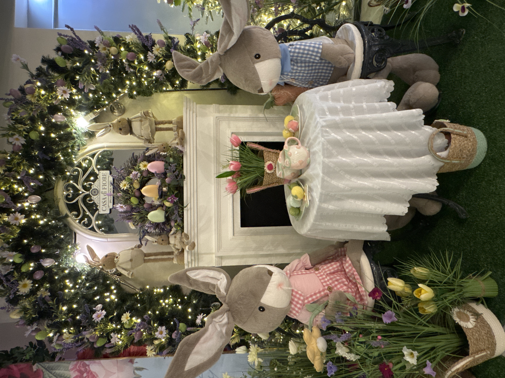 A festive Easter display includes two large stuffed rabbits. One rabbit wears a blue checkered shirt and sits on a black miniature bench at the back. The second rabbit wears a red checkered dress and sits in a similar bench in the front. A small white table sits between them, adorned with a miniature tea set, a basket of pink tulips, and decorative eggs. The backdrop features a faux fireplace decorated with an oval mirror surrounded by greenery, white flowers, lavender, and Easter eggs. Twinkling lights are intertwined within the foliage, adding to the celebratory atmosphere. Additional floral arrangements are placed around the rabbits. The setting is likely a retail display, but the location is not Kranevo, Bulgaria.