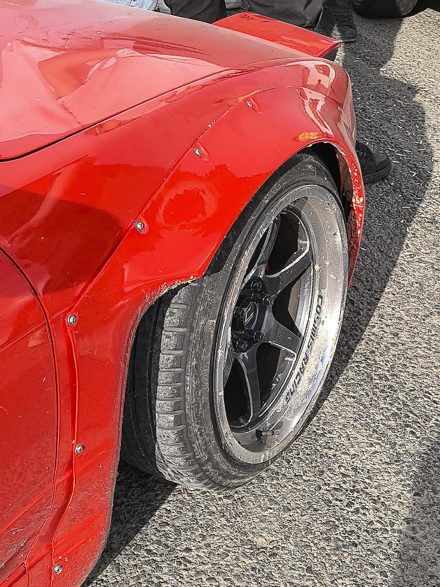 The image presents a close-up, angled view of the front right side of a bright red, custom-modified sports car, situated outdoors on an asphalt surface in Miranda de Ebro, Spain. The car features a prominent widebody front fender flare, bolted securely onto the vehicle's original bodywork. The red paint exhibits reflections, minor scuffs, and dirt, particularly around the wheel arch, which appears to have a rough, possibly cut edge.
A large, multi-spoke black wheel with a polished or silver outer lip is clearly visible. White text on the wheel lip reads "COSMIS RACING." The wide tire fitted to the wheel shows signs of wear, hinting at performance use. A tire valve stem is also discernible on the wheel.
In the background, behind the car and partially obscured by its wheel, the lower legs and feet of a person are visible, clad in dark shoes and trousers. The scene is illuminated by strong, direct sunlight, casting distinct shadows on the textured asphalt ground. The overall impression is of a modified, performance-oriented vehicle, potentially at a car show or event.