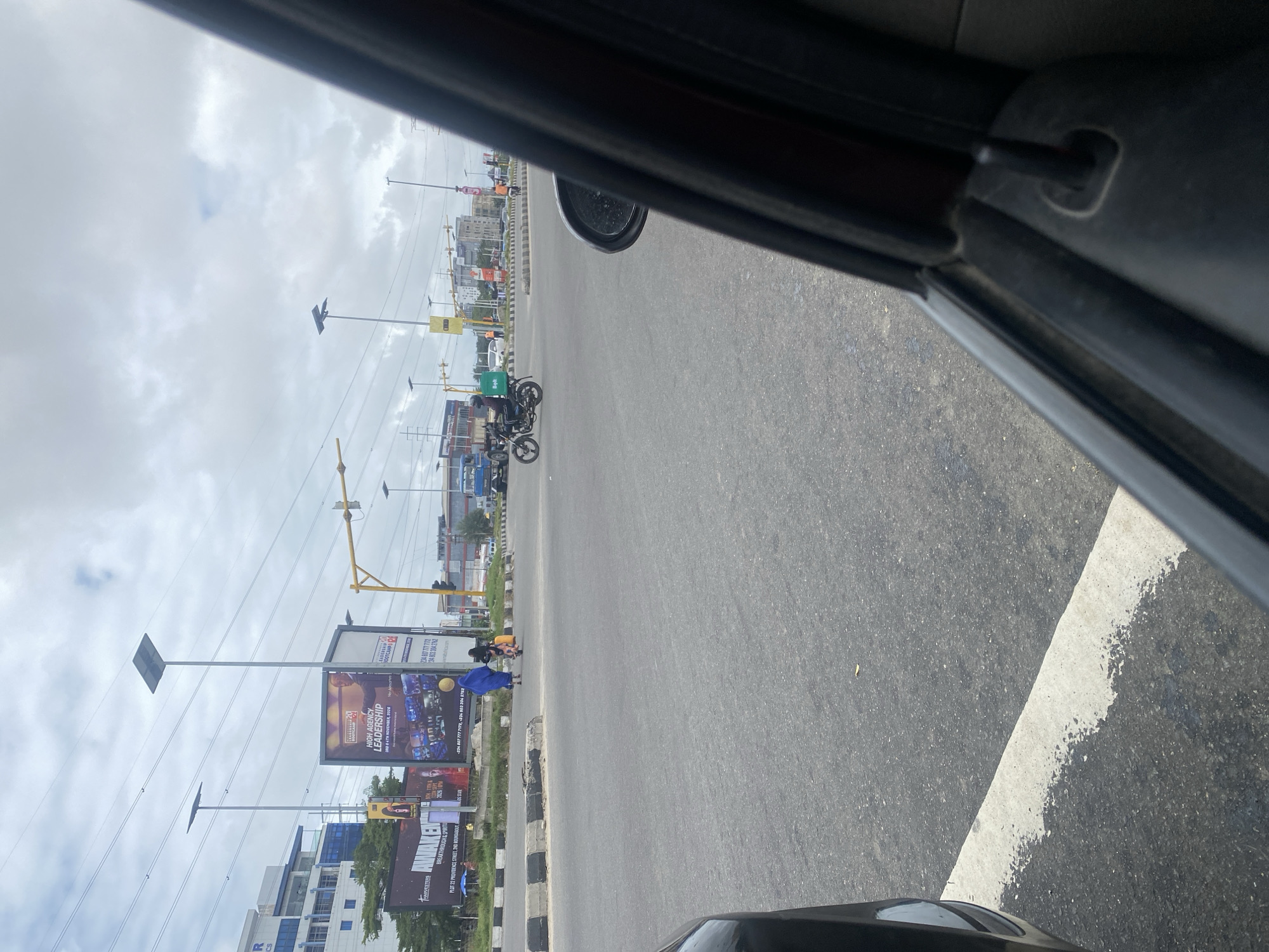 A street view in Ilado, Nigeria, captured from inside a vehicle, shows a cloudy sky over a busy intersection. The foreground is dominated by the interior of a car, with a black door frame and side mirror visible on the right. The road, marked with a white line, stretches into the distance. On the left side of the road, a large billboard with the text "AWAKEN" and "BREAKTHROUGH & SPIRITUAL" is partially visible. Further down the road, a motorcycle with a green "Bolt" delivery box is in motion, ridden by a person wearing a helmet and a dark shirt. Two individuals are seen walking across the road, one carrying a yellow bag, and the other in a blue covering. Several traffic lights, streetlights with solar panels, and buildings are scattered throughout the scene, indicating an urban environment. The overall impression is that of a typical day in a Nigerian city.