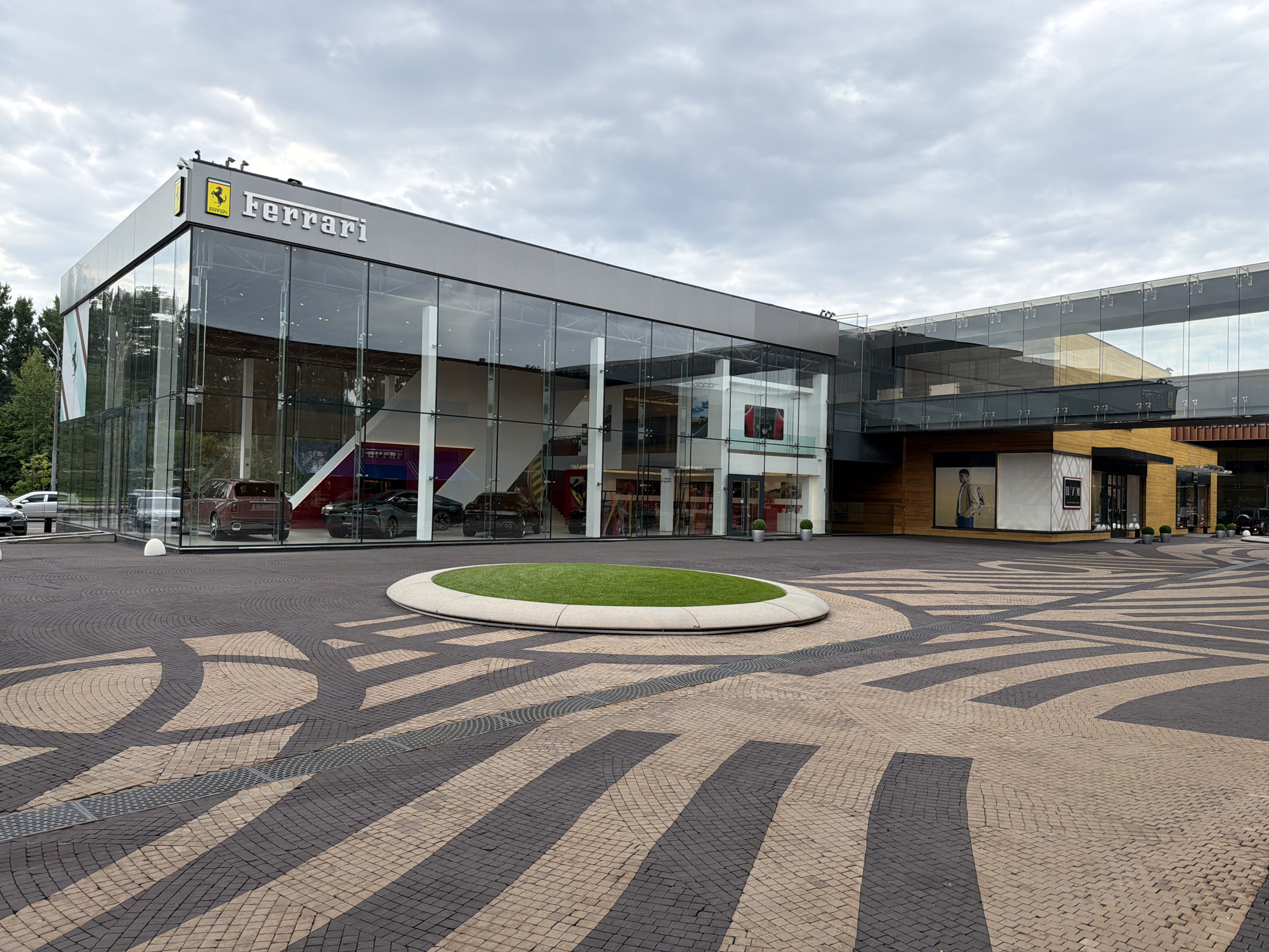 A modern Ferrari dealership with a glass facade is situated in the city of Barvikha, Russia. The building features large glass windows that offer a view of several cars displayed inside. Above the entrance, the Ferrari logo and name are prominently displayed. The surrounding area is paved with a distinctive pattern of dark and light-colored cobblestones, forming a circular design with a central patch of green grass. The sky is overcast with clouds. In the foreground, a drainage grate is visible on the cobblestone pavement.
