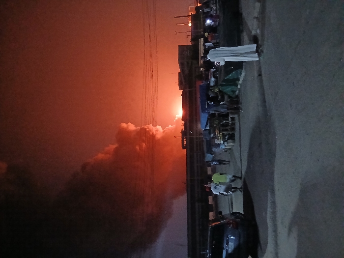 A fiery orange sunset or explosion illuminates the horizon, casting an eerie glow over a roadside scene. Stalls line the edge of a road, populated by blurry figures of people. A man in white robes stands out, adding a touch of solemnity. A car sits parked nearby, its dark shape a stark contrast to the vibrant sky. Power lines extend from the middle of the image vertically, emphasizing the industrial nature of the surrounding area.