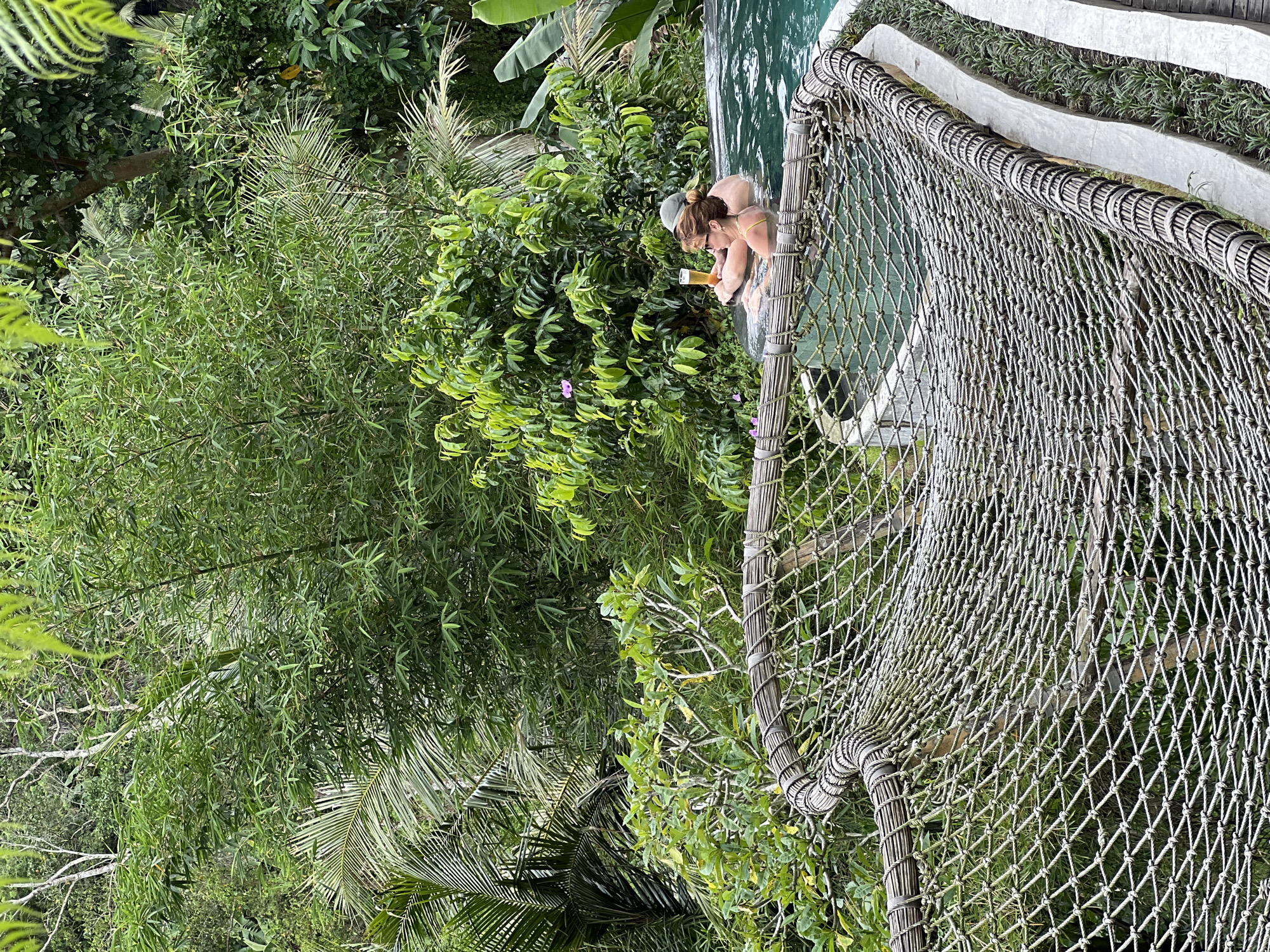 A high-angle shot shows a couple in a pool, likely at a resort or villa, in Tegallalang, Indonesia. The pool is surrounded by lush green vegetation, including bamboo, various trees, and tropical plants. A rope net structure partially covers the foreground and extends over the pool area. One person, presumably a woman, wears a light-colored top and drinks from a glass or can. A man is next to her in the pool. The architecture incorporates natural materials, and the general environment appears tropical and serene.
