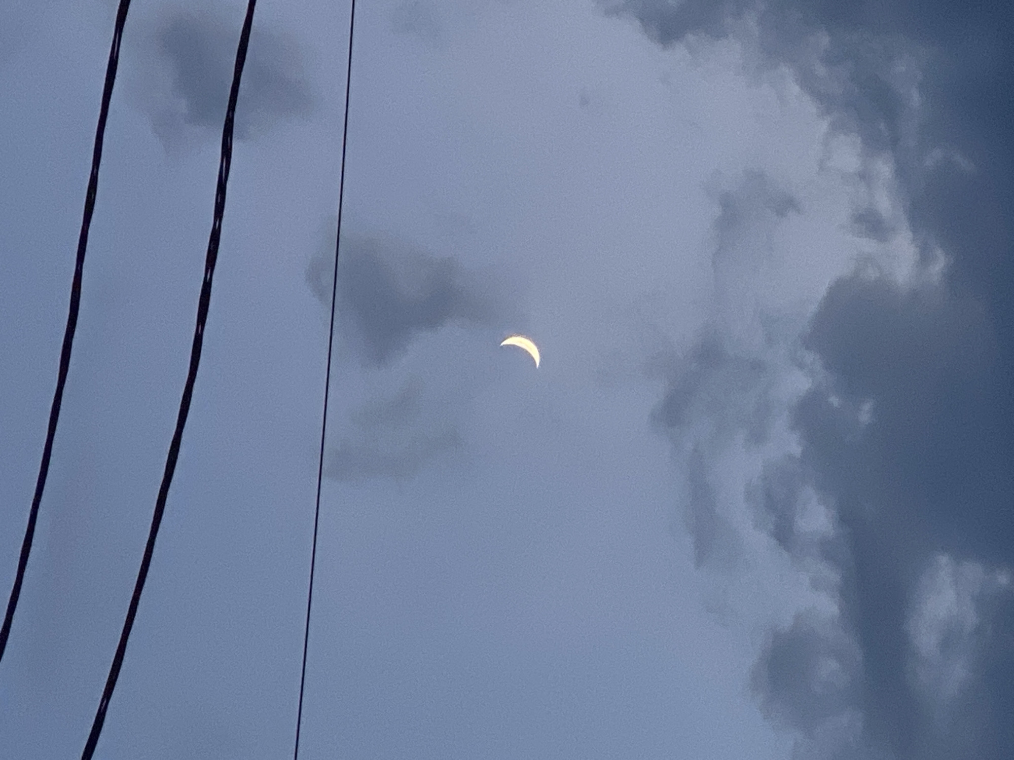 The image depicts a crescent moon in a dusky, cloudy sky, with power lines running horizontally across the upper portion of the frame. The sky is a muted blue-gray, with scattered dark clouds. The moon is a thin sliver of light, positioned slightly to the left of the center. The power lines are thick and dark, casting no visible shadows. The scene is captured from a low angle, looking upwards. There are no people or specific landmarks visible that would indicate the location within Vysokoye, Russia, beyond the general context provided. The overall impression is one of a serene, twilight sky.