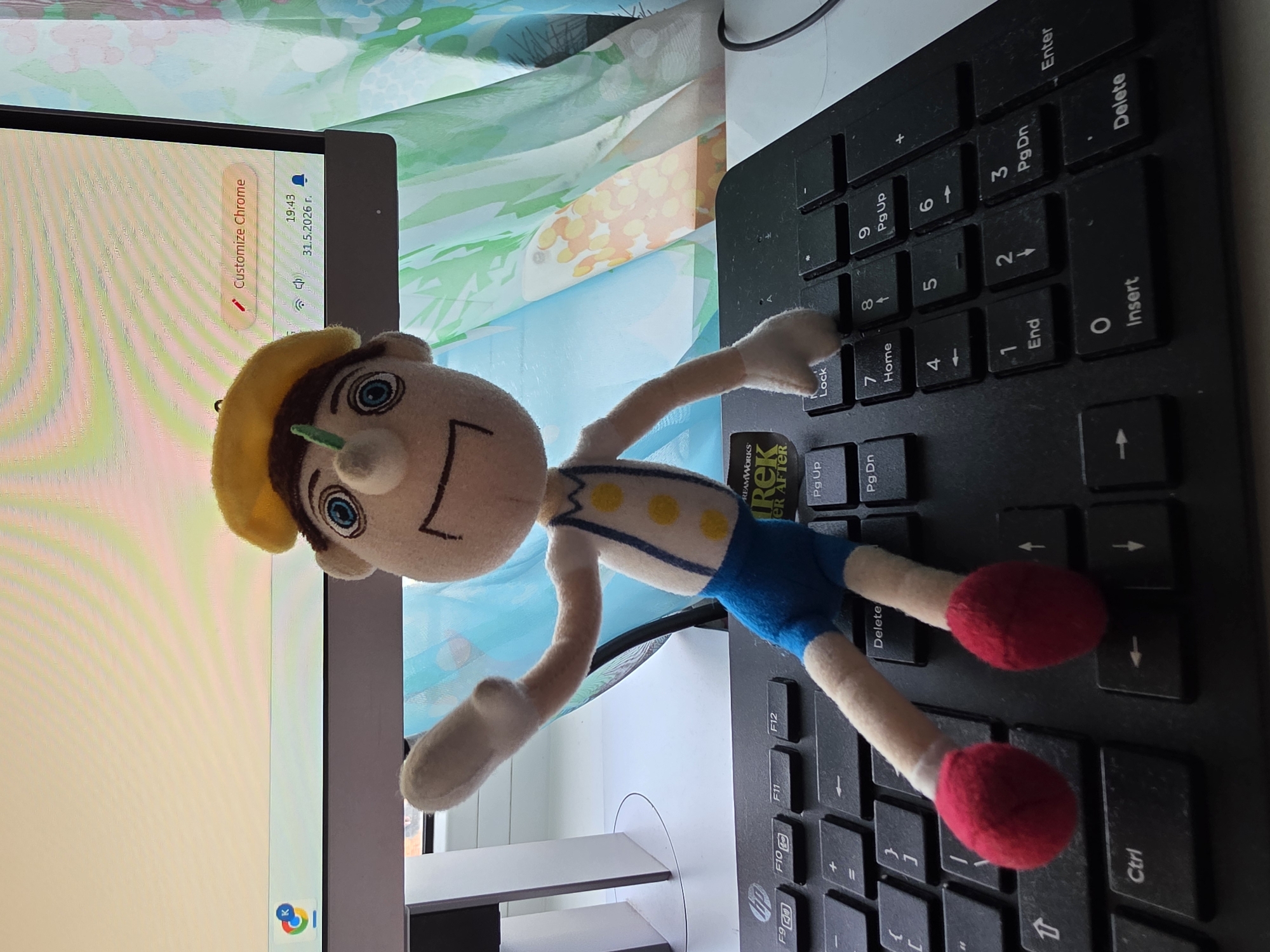 A plush toy of Pinocchio is lying on a black computer keyboard. The keyboard is positioned next to a computer monitor, which displays a Chrome browser window, timestamped 19:43, 31.5.2026 г. The scene is indoors.