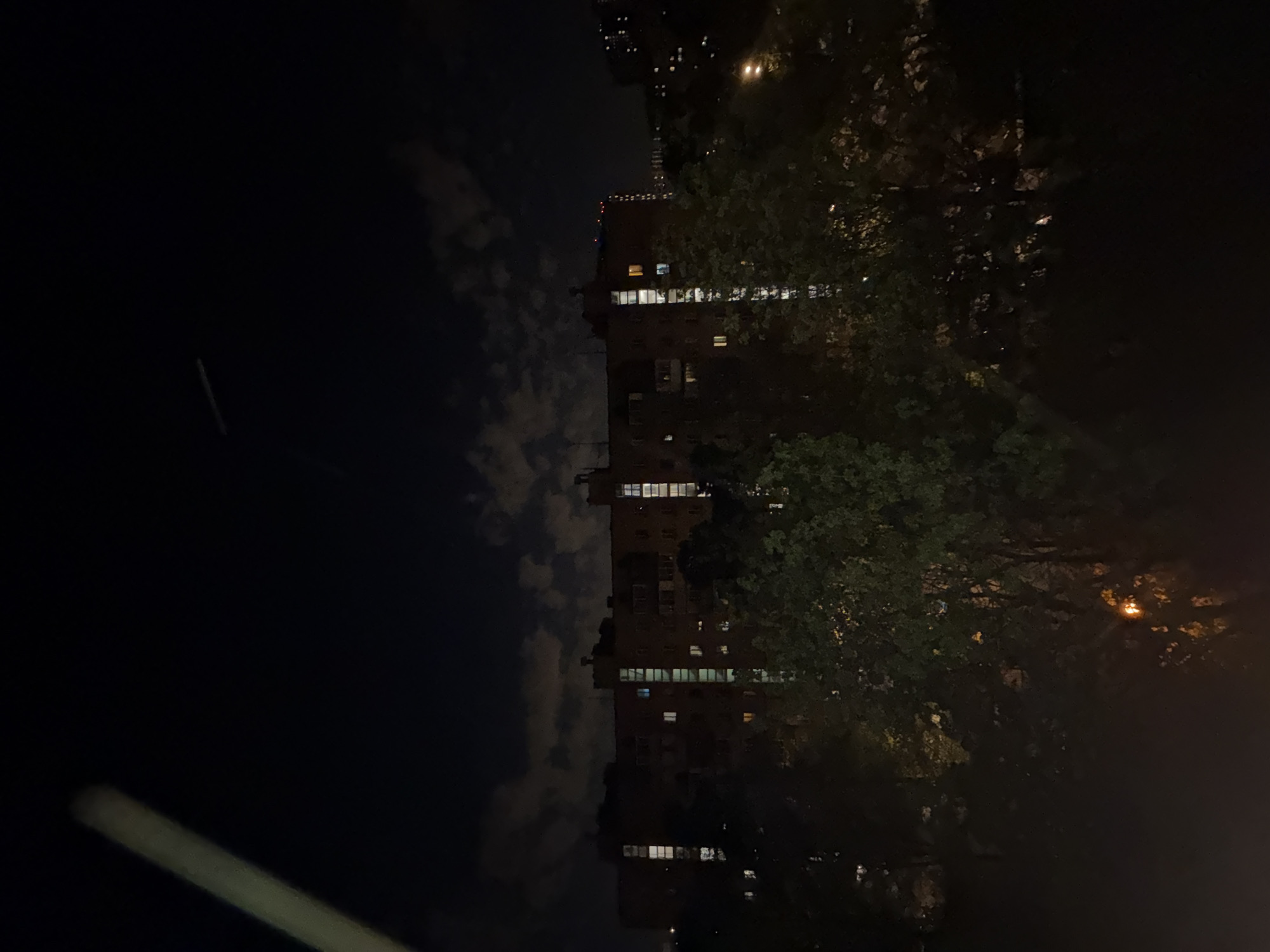 The image shows a nighttime scene in what appears to be an urban environment. A tall building with illuminated windows is visible, partially obscured by trees on the right. Clouds are present in the sky on the left side of the image. A light streak is seen in the lower-left corner, likely from a passing vehicle or light source. The overall atmosphere is dark, with limited lighting. Without additional information or details included in the photo, it's not possible to pinpoint the precise location within Mytishchi, Russia.