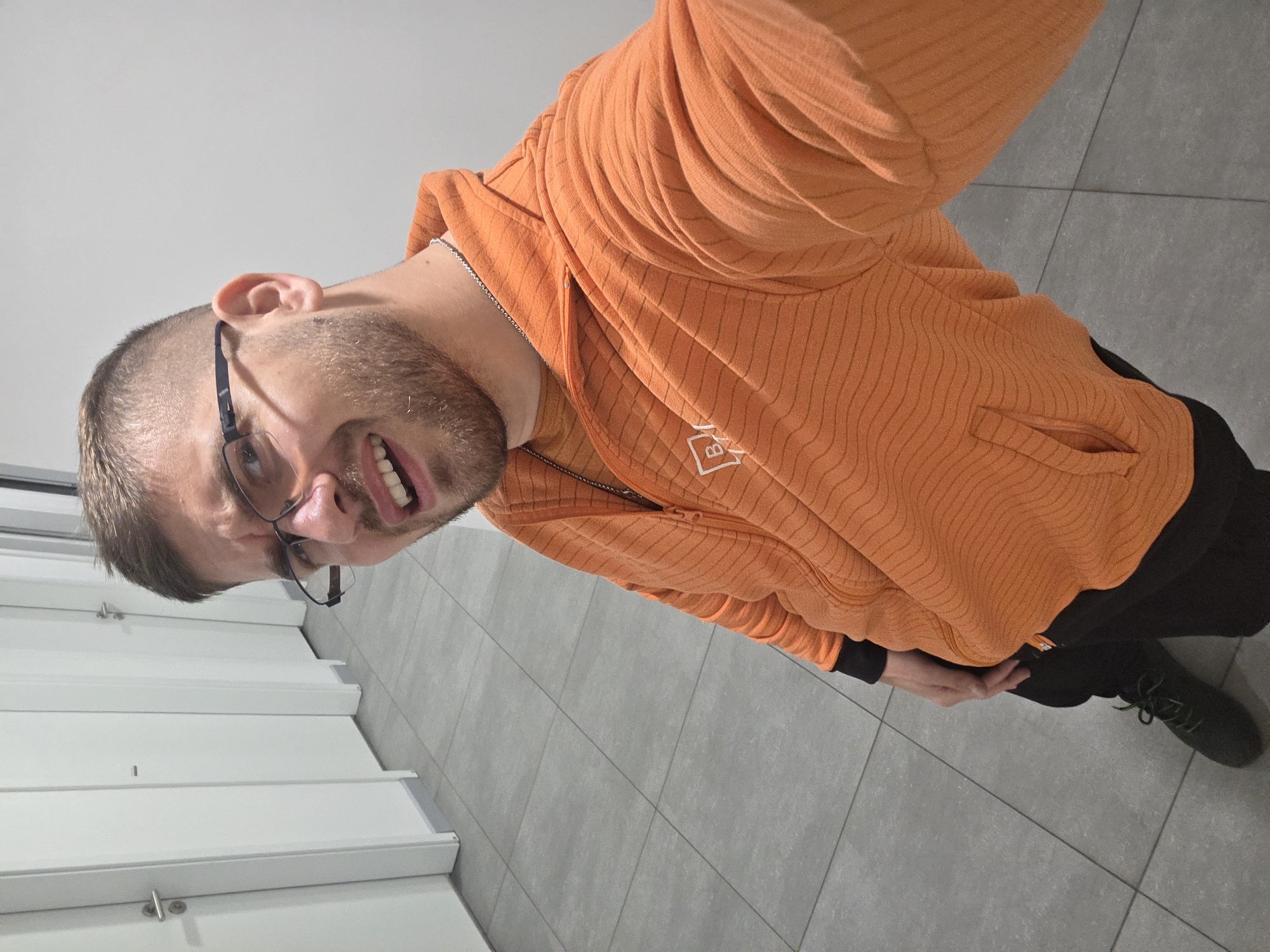 A man with short, light brown hair, a beard, and glasses is taking a selfie. He is wearing an orange hooded sweatshirt with black sleeves and pants. He is standing on gray tiled flooring, next to what appears to be white wall panels. The location may be in the city of Plovdiv, Bulgaria, although no specific landmarks indicating this are directly visible in the image.