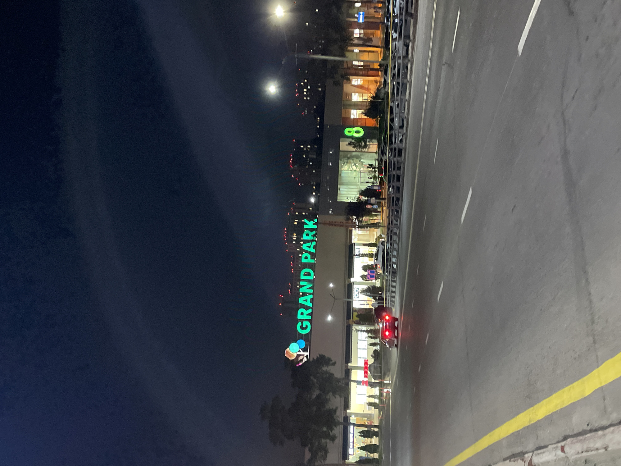 This is a night-time, eye-level, medium shot of the Grand Park shopping mall, located on Al-Farabi Avenue in Almaty, Kazakhstan. The prominent green neon sign "GRAND PARK" is visible across the top of the building. To the right of the sign, a large neon green number "8" is illuminated. The building features large glass windows and entrances, with some interior lights visible. A row of trees and decorative shrubs line the exterior of the mall. A street with multiple lanes of traffic runs in front of the mall, separated by a barrier. A single red car with illuminated taillights is driving away from the camera on the street. Several other cars are parked in a lot behind the barrier. The sky is dark and cloudy, with scattered lights from distant buildings visible in the background. The overall scene depicts a commercial area at night.