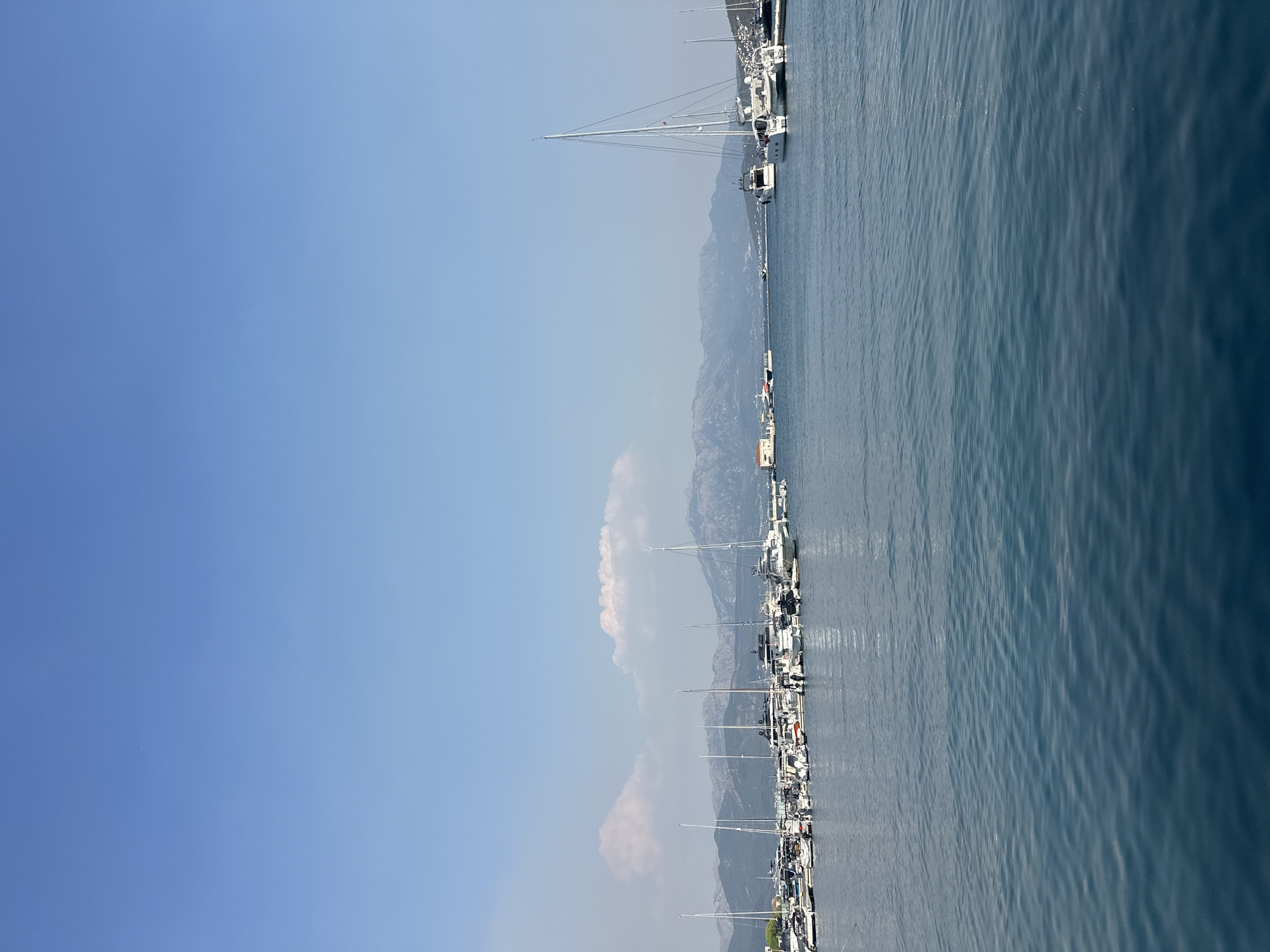 The image shows a wide shot of a harbor in Herceg Novi, Montenegro, under a clear blue sky. The water in the foreground is a deep blue with gentle ripples. In the middle ground, a large number of yachts and smaller boats are moored at docks. These boats vary in size and design, with many featuring white hulls and masts. Beyond the harbor, a range of mountains forms the background, with hazy peaks suggesting distance. A few wispy clouds are scattered across the sky. On the far right of the harbor, a building complex with white structures is visible, and a few boats are also seen sailing in the distance.