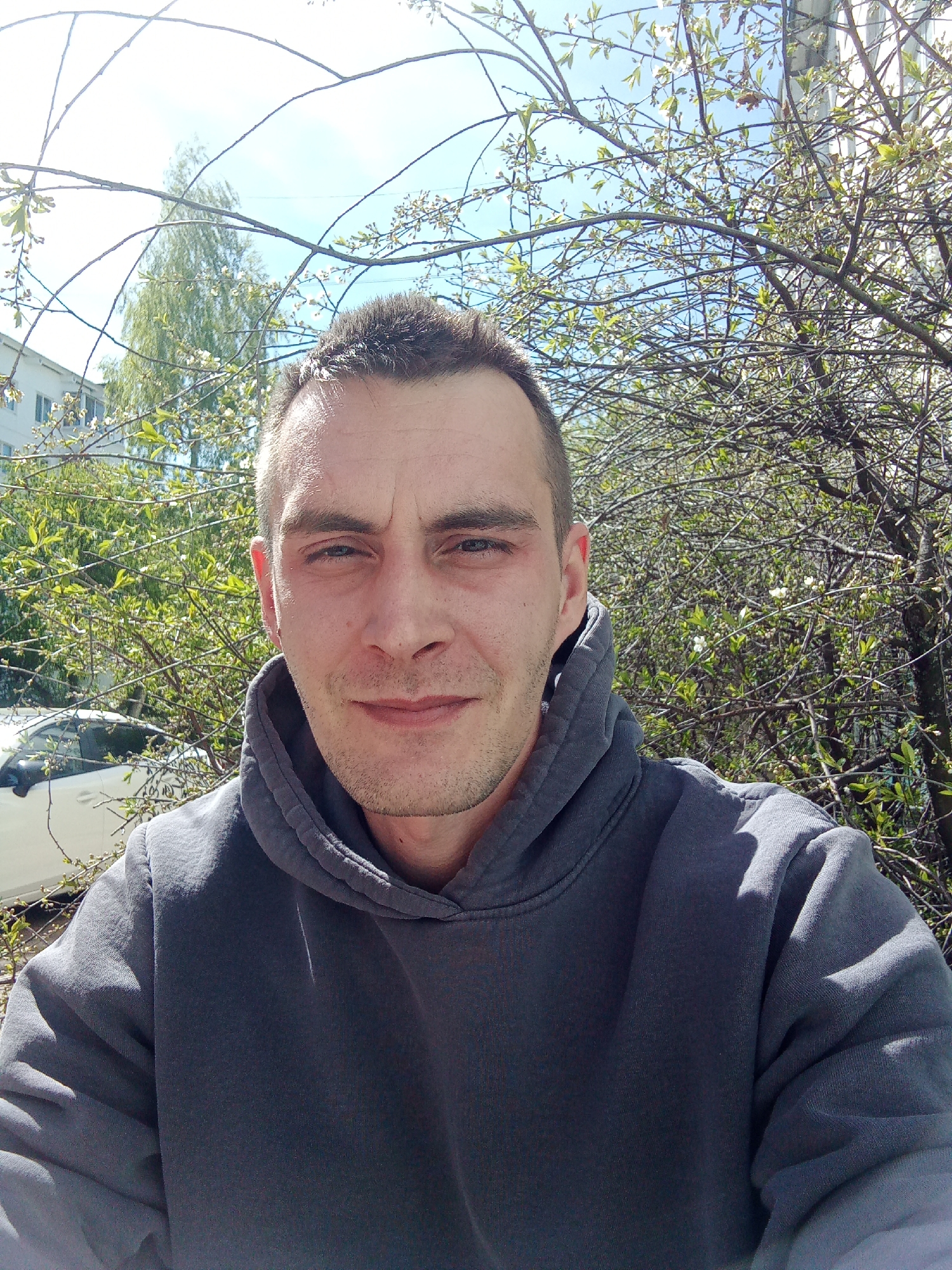 The image shows a man in a gray hoodie taking a selfie outdoors. He has short dark hair, fair skin, and blue eyes. He is looking directly at the camera with a slight smile.
The background is filled with trees, some of which have white blossoms. A white car is partially visible in the lower left of the frame, along with the side of a multi-story building with windows. The sky is light blue and sunny.
The photograph appears to have been taken in the city of Chaikovsky, Russia.