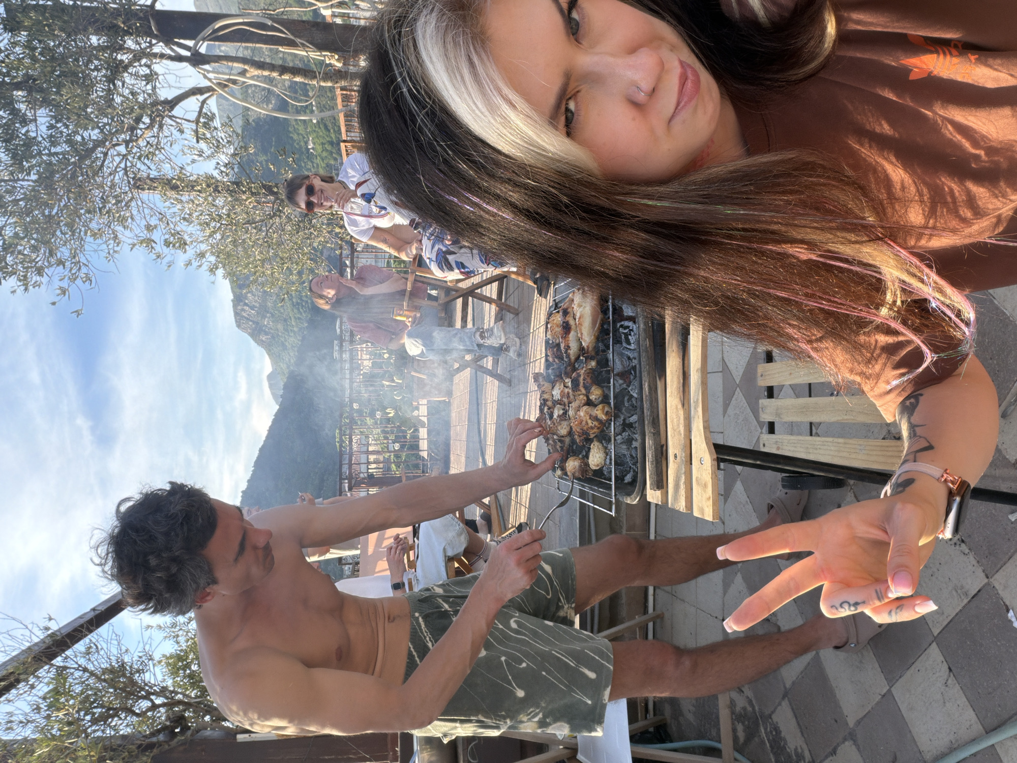 A group of people are gathered outdoors, presumably enjoying a barbecue. A woman with dark hair, featuring a noticeable white streak, is positioned in the foreground, possibly taking a selfie. She is wearing a brown t-shirt and shows a peace sign with her hand. Her hair also has some colored strands in it. A man, shirtless and wearing green and white patterned shorts, is standing near a grill, using a fork to manage food that appears to be some kind of poultry grilling, on the grill. Smoke from the grill rises behind him. In the background, seated people are visible. Beyond them is a landscape featuring a mountain under a blue sky, partially obscured by the grill's smoke. The location appears to be a patio or terrace. Given the context of the request, it is likely that the location is within or near Budva, Montenegro, based on the prompt's explicit reference to the city.