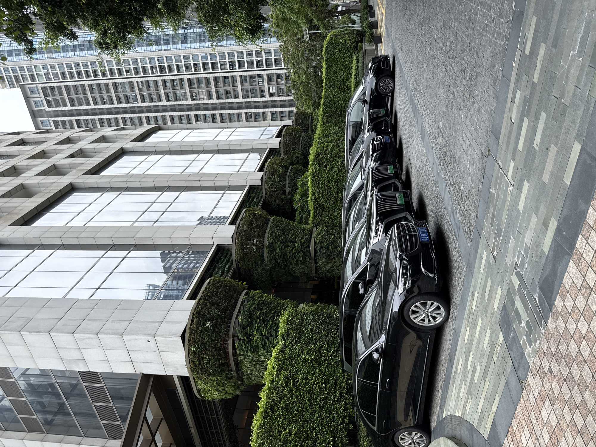 A line of black luxury vehicles, including sedans and vans, are parked on a cobblestone driveway in front of a modern building. The building features a facade of large glass windows and is adorned with vertical gardens on its exterior. In the background, taller residential buildings are visible. The scene appears to be in a commercial or business district, with lush greenery contributing to the urban landscape. The overall impression is one of sophistication and urban development.