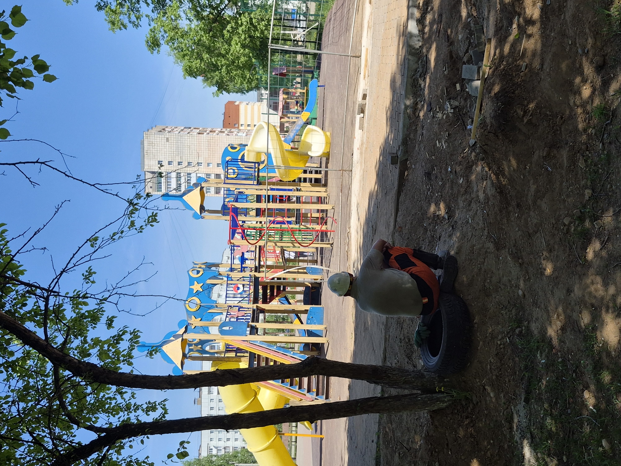 A person sits on a tire near a playground in the city of Unknown, Unknown. The playground has slides, climbing structures, and other play equipment, with apartment buildings in the background. The individual is wearing a light-colored shirt, dark pants, and a light-colored hard hat. The surrounding area is partly shaded with trees.