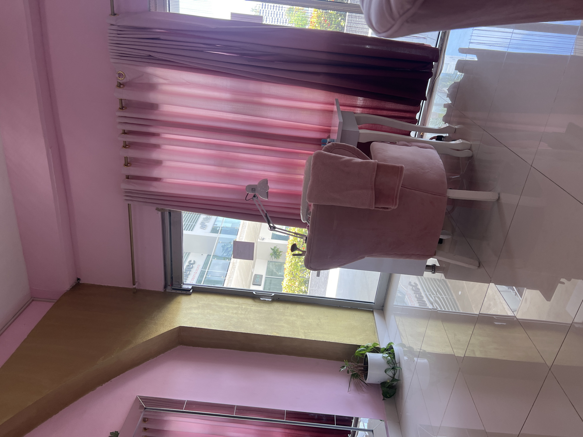 The image depicts the interior of a nail salon in Cancún, Mexico. The room is decorated in shades of pink and gold, with plush pink seating and ornate white furniture. Pink gradient curtains hang from a curtain rod, covering a large window that offers a view of the city and surrounding greenery. A white desk with a lamp and a few brushes sits near a pink armchair. The floor is made of large, reflective white tiles. A potted plant is visible in the foreground. The overall ambiance suggests a luxurious and feminine space designed for beauty treatments.
