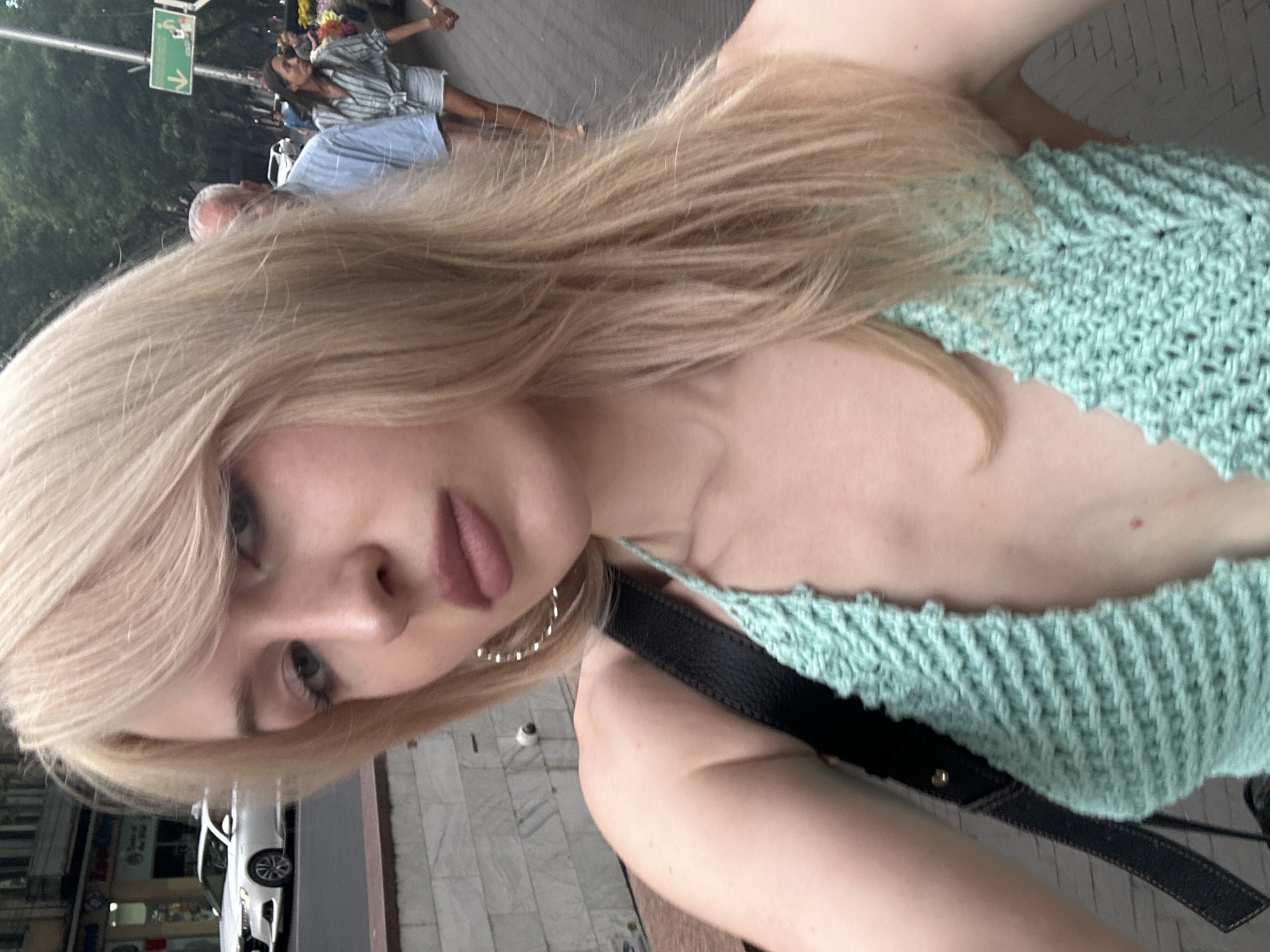 A young woman with blonde hair takes a selfie while walking on a city sidewalk. She is wearing a light blue, crocheted halter top and large hoop earrings. In the background, a white car is parked on the street, and other people are walking along the sidewalk, some with flower stalls visible. The scene appears to be in Tbilisi, Georgia, as indicated by the architecture and street signage.