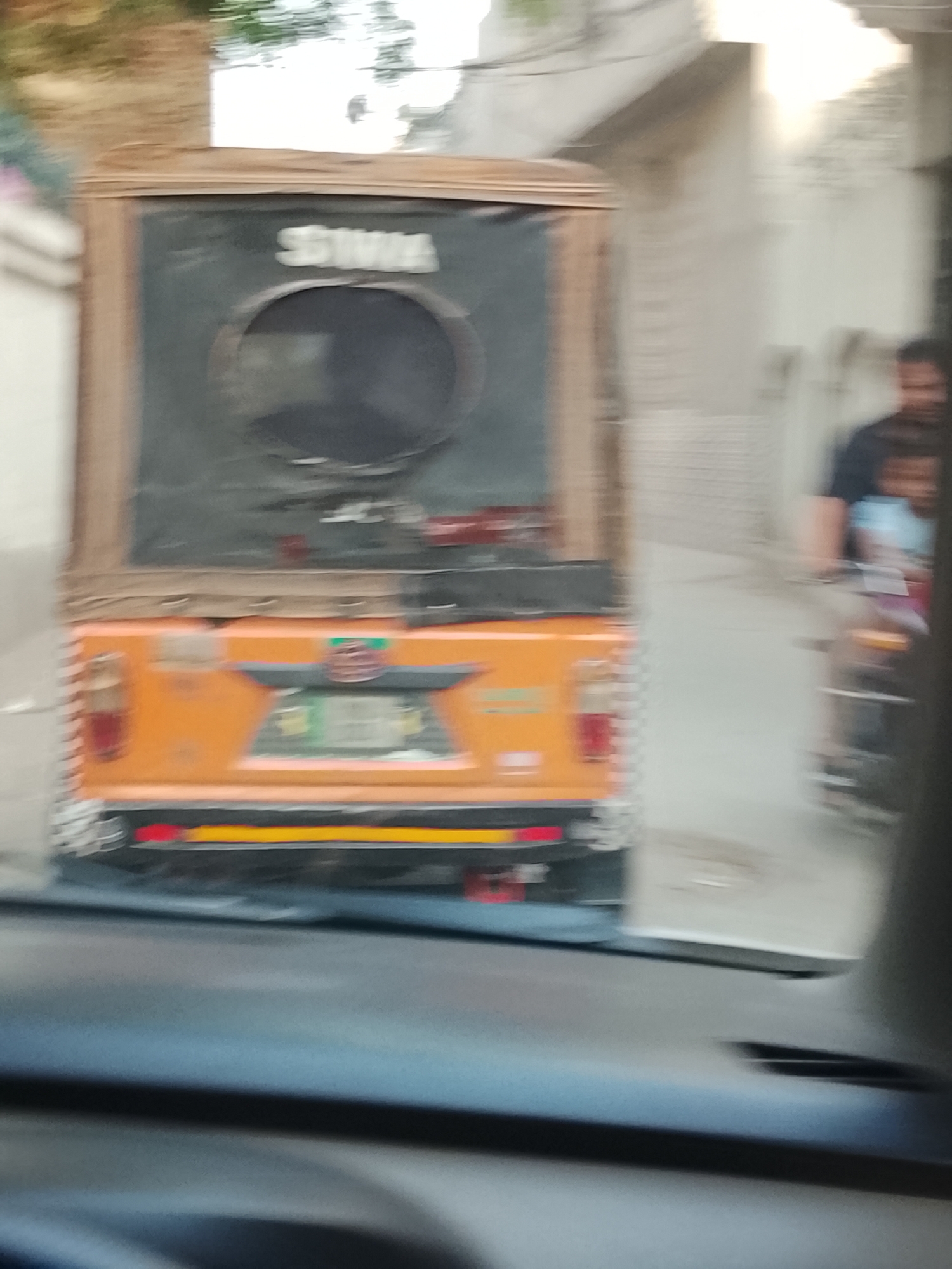 The image depicts the rear of an auto-rickshaw in a blur, likely taken from inside a car following it. The rickshaw is orange with a black canvas covering the back. There's a circular window in the canvas. On the orange section, there are tail lights and a license plate that is blurred. Behind the rickshaw, a man and child appear to be on a motorbike. The background includes buildings, suggesting an urban environment, which could be Nasheman Colony, Pakistan. The image is blurred, suggesting movement.
