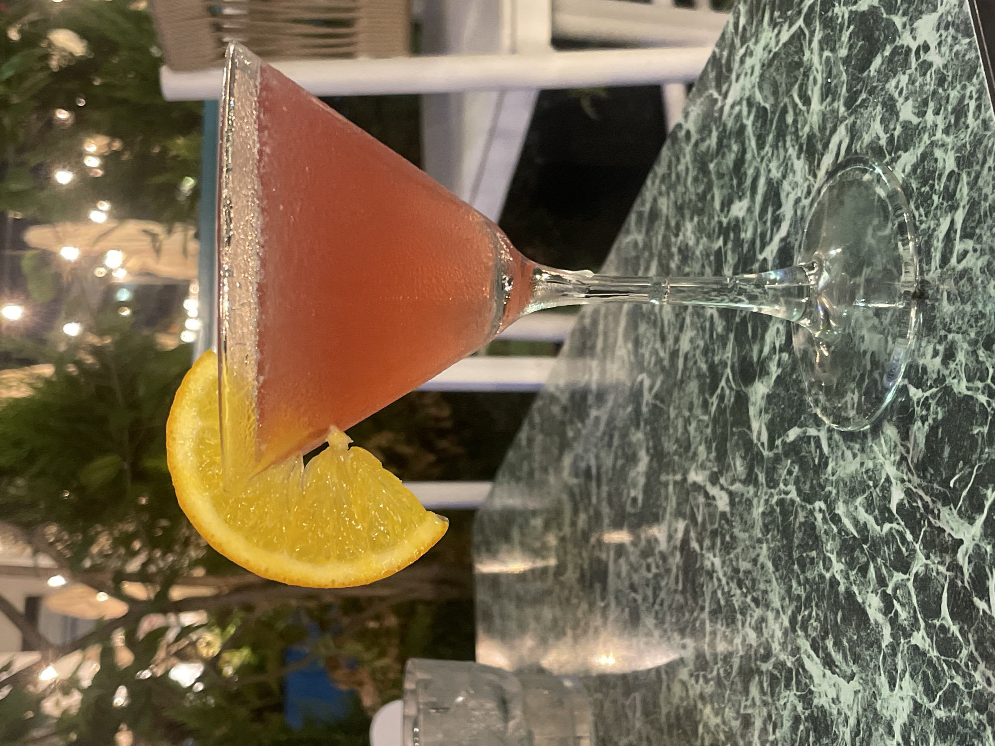 A close-up shot shows a pink cocktail in a martini glass, garnished with an orange slice. The cocktail is on a dark green marble table. In the background, blurry lights and greenery suggest an outdoor evening setting, possibly a restaurant or bar. The location is identified as Herzliya, Israel.
