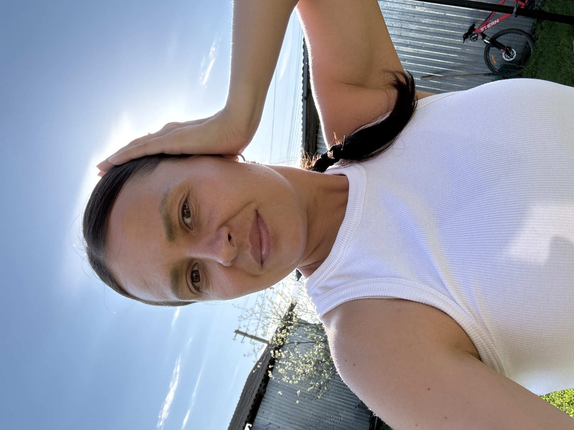 A woman with dark hair pulled back into a braid and wearing a white tank top is holding her hand to her forehead, possibly shielding her eyes from the sun. The background shows a corrugated metal fence, grass, a bicycle, a tree, and a blue sky with some clouds. The location cannot be confirmed as Чумна, Russia from this image.