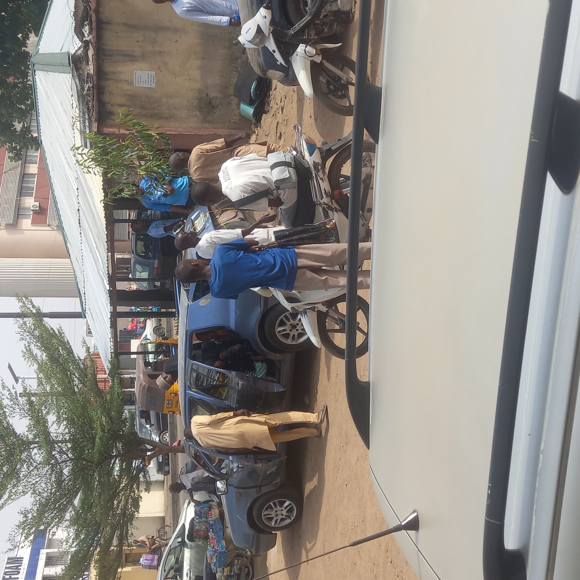 The image captures a street scene in what appears to be a bustling urban environment in Dadi Riba, Nigeria. Several individuals are present, some standing near parked vehicles, including cars and motorcycles. One person is seen entering or exiting a blue car with its door open. Other vehicles, including a yellow auto-rickshaw (tuk-tuk), are visible in the background, suggesting a busy street. The setting includes buildings with metal roofing and concrete walls. A tree with green foliage is also present, contributing to the urban landscape. There is a business visible in the far background (FOAM) suggesting a commercial environment. The overall scene depicts everyday life and activity in this part of the city.