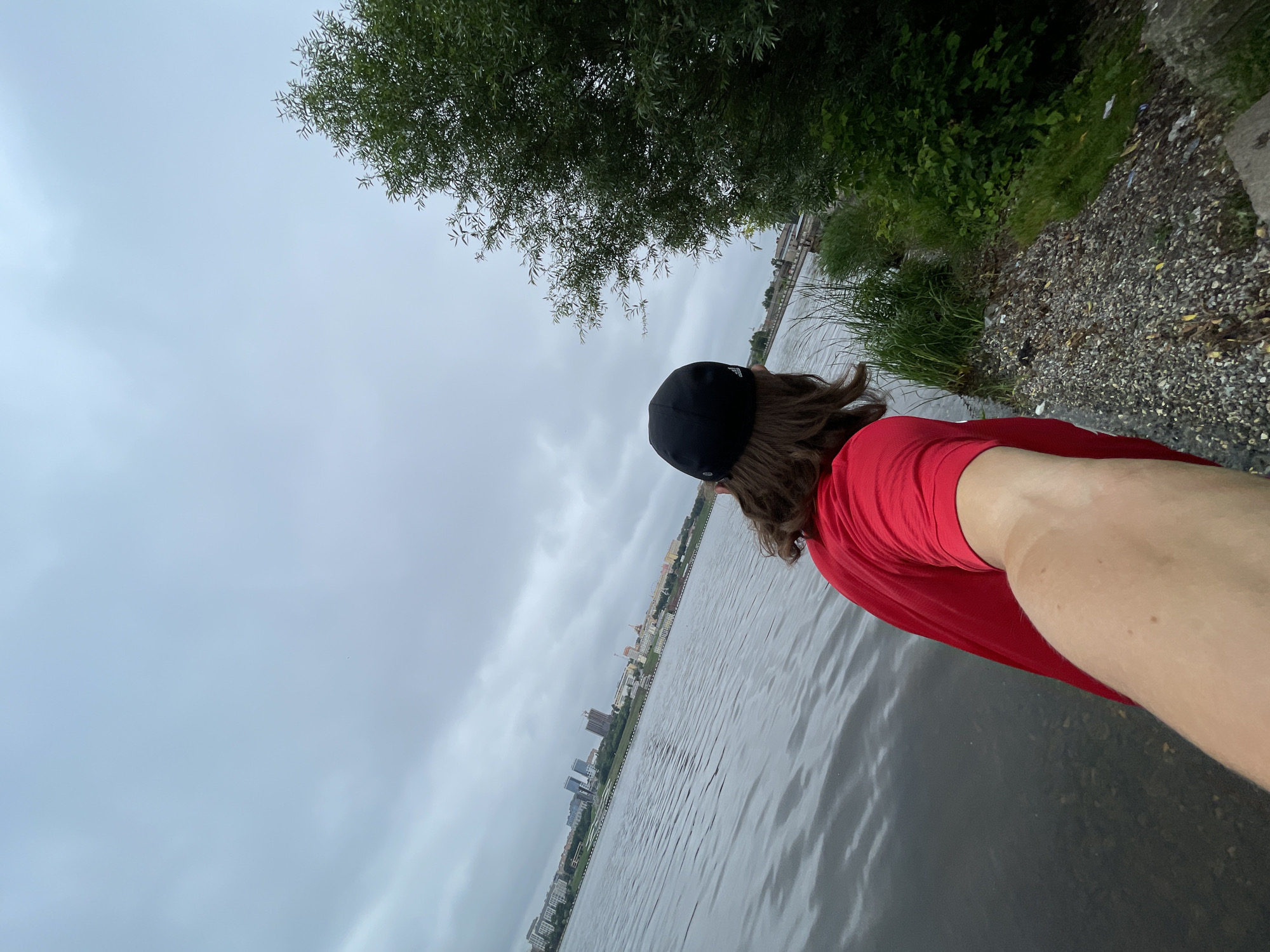 A person is seen from behind, wearing a red t-shirt and a black baseball cap, standing at the edge of a body of water. The person's right arm is extended towards the camera, as if taking a selfie. In the background, across the water, a city skyline is visible with buildings of various heights. The sky is overcast with grey clouds. To the right of the person, there is a dense green tree and overgrown grass. The ground in the foreground is covered with small stones and debris. The water appears calm with ripples.