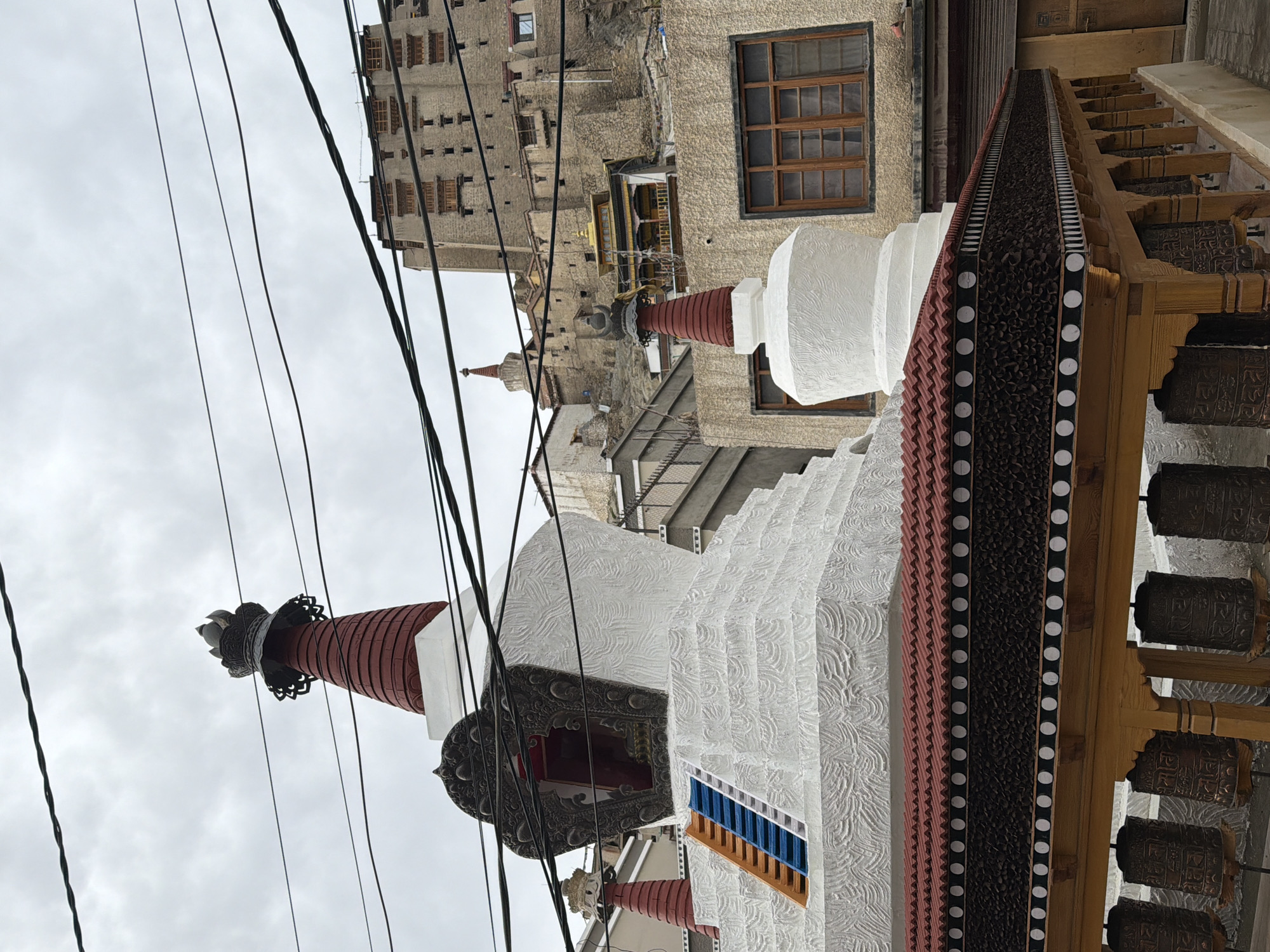 The image shows a view of the Leh Palace and a Buddhist stupa in Leh, India. The Leh Palace is a prominent landmark, a nine-story royal palace on a hilltop overlooking the town. In the foreground, a white stupa with a red spire stands next to a wooden structure adorned with prayer wheels. The sky is overcast, with power lines crisscrossing the frame. The architecture suggests a blend of traditional Tibetan Buddhist and Ladakhi styles.