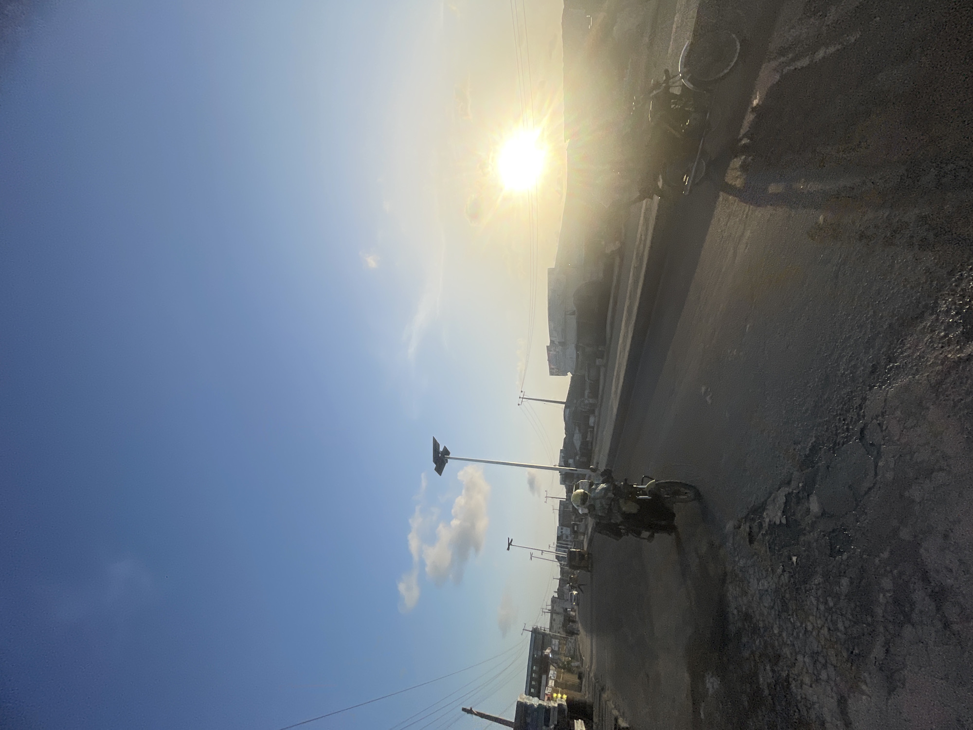 The image shows a road in Ado, Nigeria, on a sunny day. The sun is high in the sky, partially obscured by haze. Along the road, there are streetlights and power poles. A motorcycle is moving along the road. The road surface appears cracked and worn. Buildings are visible in the background.
