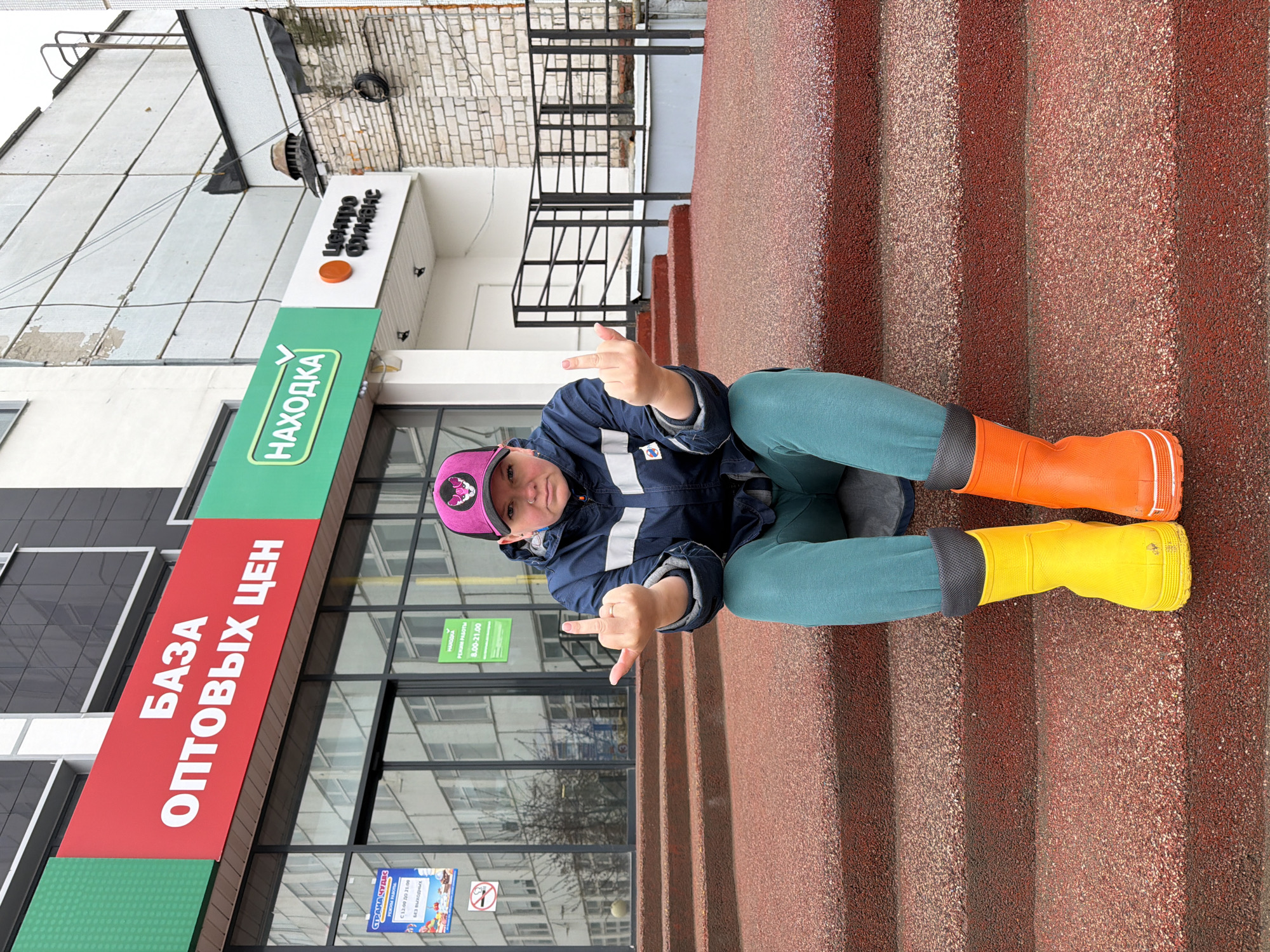 A person wearing a blue jacket, green pants, a pink cap, and mismatched yellow and orange boots is sitting on red stairs in front of a building. The person is holding up both hands, each with the middle finger extended. The building has signs in Russian, including "НАХОДКА" (NAKHODKA), "БАЗА ОПТОВЫХ ЦЕН" (WHOLESALE PRICE BASE), and "центро Финанс" (Centro Finance). A sign for a business called "Страна Чудес" (Country of Wonders) with stated business hours is also visible on the building. Based on the signs and linguistic details, the location is likely in Chaikovsky, Russia.