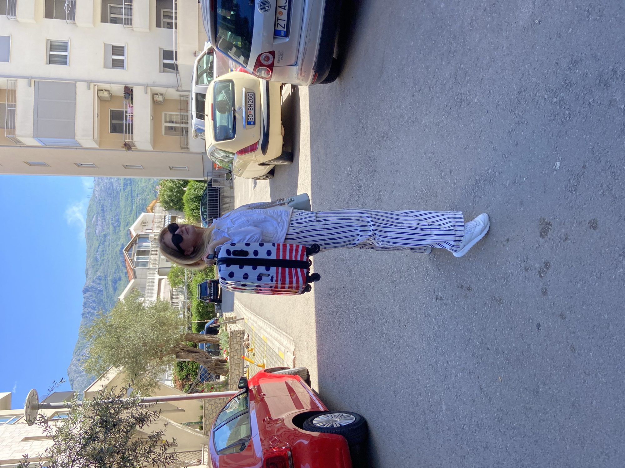 The image captures a woman standing on an asphalt street in Budva, Montenegro, under a clear, sunny sky, likely during midday or early afternoon. She is dressed in a white short-sleeved shirt, white and navy blue striped wide-leg trousers, and white sneakers, with sunglasses resting on her face. A light blue handbag is visible on her shoulder. She is holding a distinctive suitcase with a white background featuring black polka dots on the top half and red and white vertical stripes on the bottom.
Around her, the scene depicts a residential area with parked cars and buildings. To her immediate right, a red car is parked. Behind her, a cream/yellow car with the license plate "BD BH 266" is visible, along with a silver/grey car whose license plate partially shows "ZT AJ" and the country code "MNE" (for Montenegro). In the background, multi-story residential buildings with balconies are visible, interspersed with green trees. Beyond the buildings, large, rugged mountains rise under a bright blue sky, characteristic of the Montenegrin landscape. The woman appears to be pausing or waiting, perhaps in transit with her luggage.