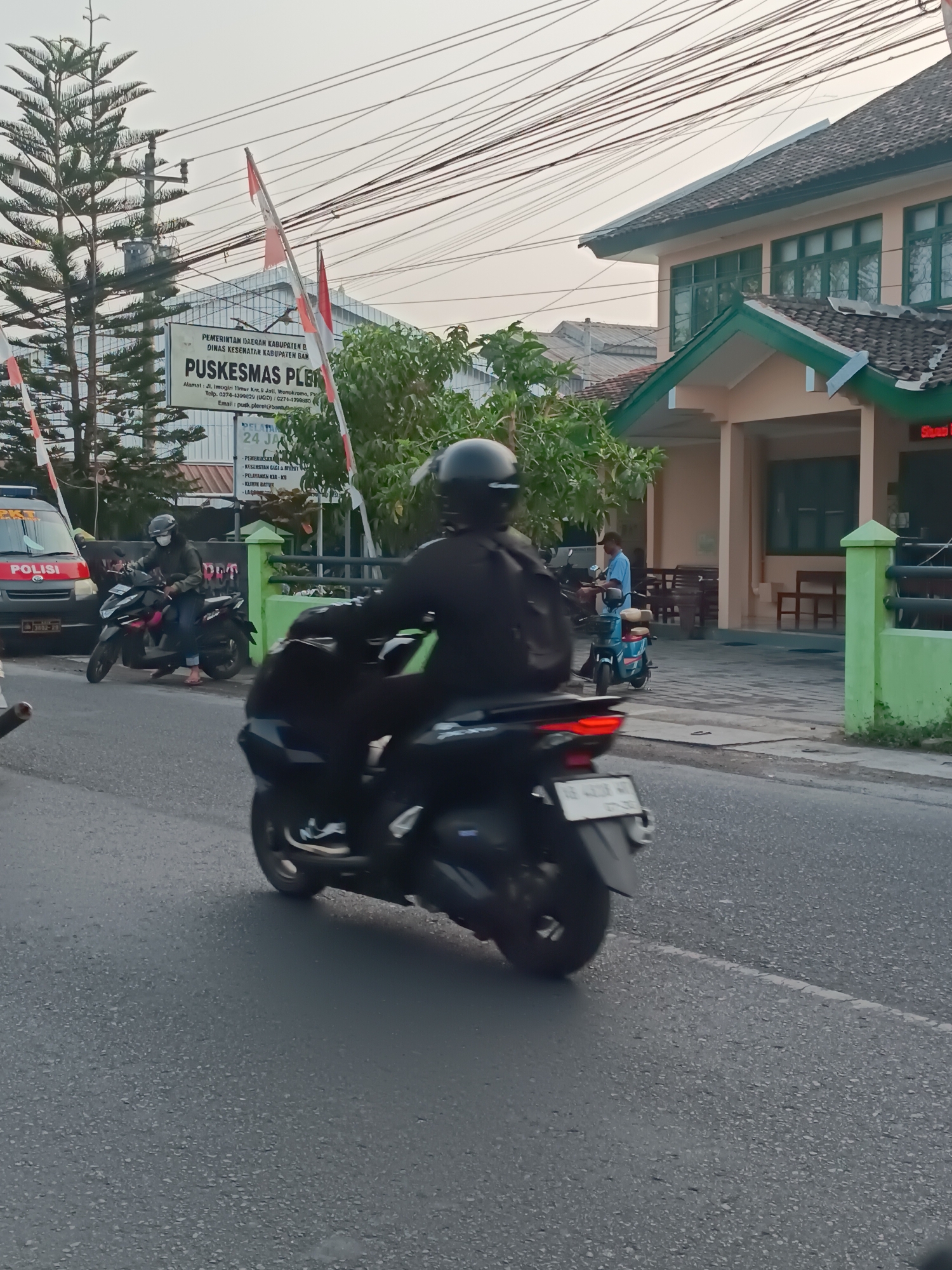 The image depicts a scene on a street in Pleret, Indonesia, likely in front of a public health center (Puskesmas). In the foreground, a person wearing a helmet and a dark jacket is riding a black motorcycle, which is blurred due to motion. Another person on a black motorcycle is visible to the left, also wearing a helmet and a mask, and is stopped near a police vehicle. The police vehicle is white with red and blue markings, and the word "POLISI" is visible on its side. Behind the motorcycles, a building with a green trim and tiled roof is present. A sign on the building reads "PUSKESMAS PLERET". Several trees and power lines are also visible in the background, along with more flags and what appears to be a barrier. The overall environment suggests an urban or semi-urban setting, possibly during daylight hours with overcast skies.