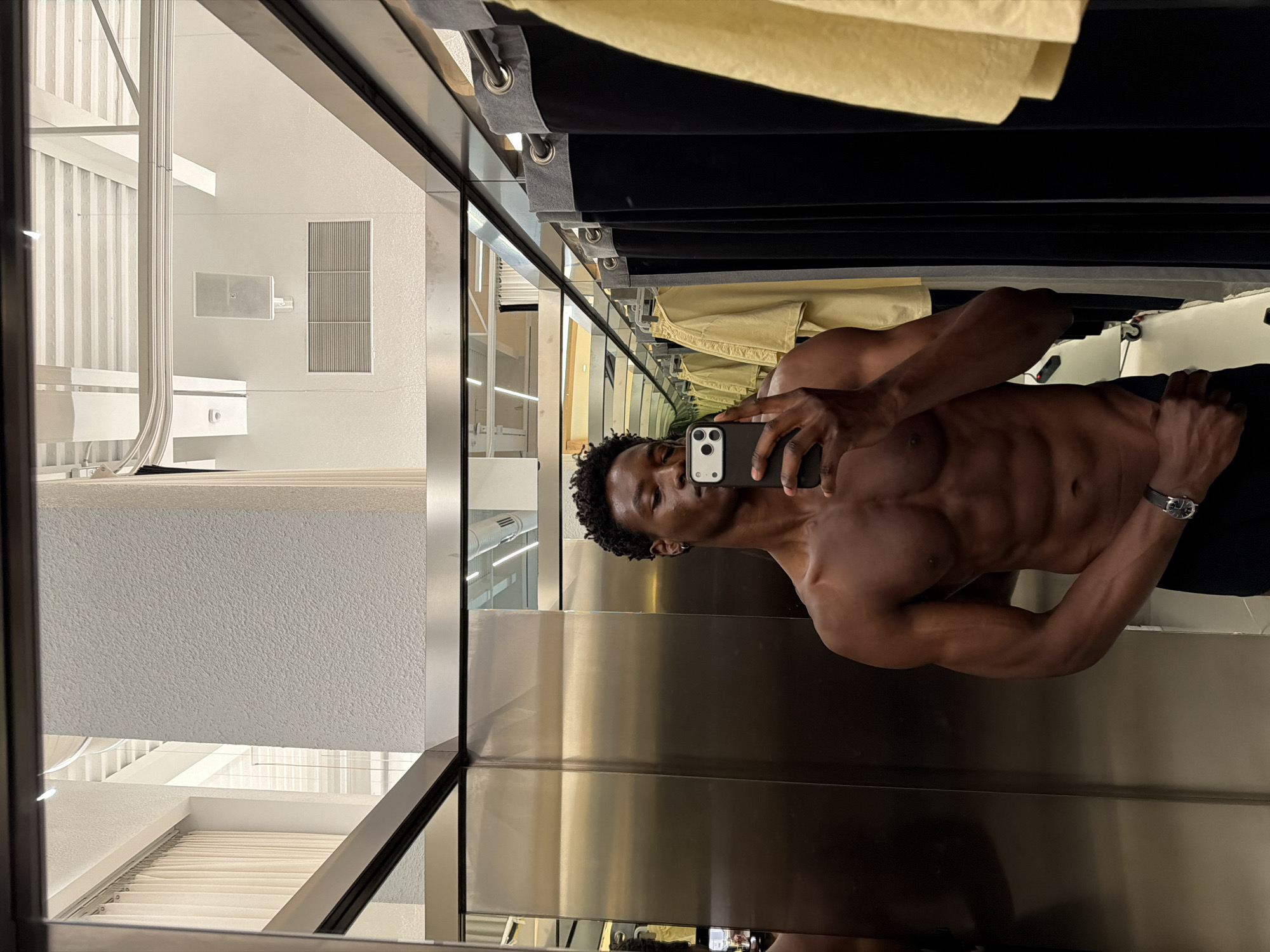 A shirtless, muscular Black man is taking a selfie in a dressing room. He is holding a phone with a triple-lens camera in his right hand, positioned to capture his upper torso and face. He is wearing a wristwatch on his left wrist and dark-colored pants. The dressing room has shiny, vertical metallic panels. Above him are clothes hanging on a rack. The reflection in the mirror shows a glimpse of what appears to be a store interior with white fixtures and geometric design elements. The location, Fibracolor, Spain, cannot be confirmed from the details of the image alone.