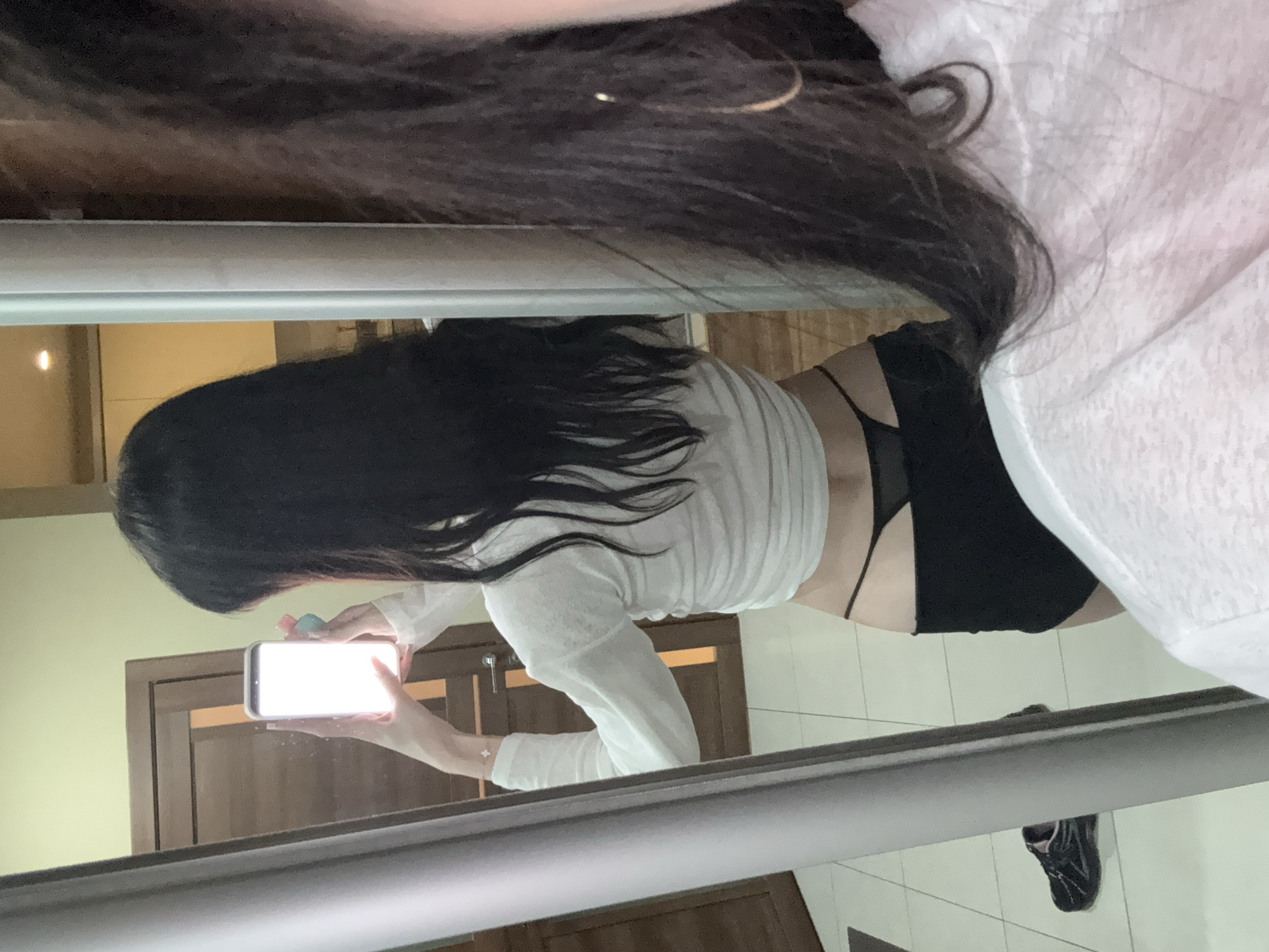 The image shows a person taking a mirror selfie. The person is facing away from the camera, holding a smartphone in their right hand, which is illuminated by its screen. They have long, dark hair and are wearing a white long-sleeved top and black shorts. The background appears to be an indoor setting, possibly a bedroom or dressing area, with a door and tiled flooring visible. A pair of black athletic shoes is on the floor. The reflection in the mirror also shows part of another person's face and shoulder on the right side of the image, also wearing a white garment. The context of Khimki, Russia, is not evident from the visual information in the image.