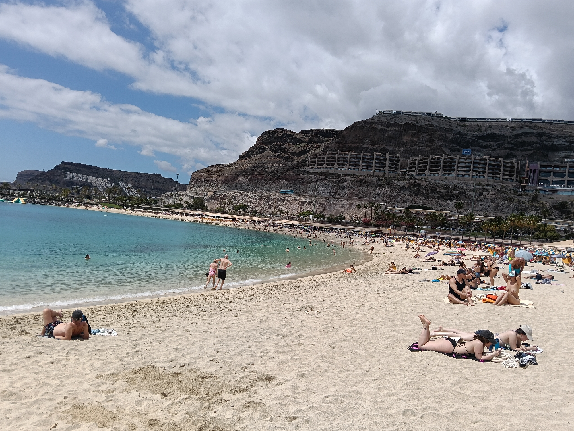 This wide-angle daytime photograph captures a vibrant beach scene in Mogán, Spain, featuring a sandy shore, clear turquoise waters, and imposing cliffs lined with terraced buildings under a partly cloudy sky.
In the foreground, the light-colored sand is dotted with numerous beachgoers engaged in leisure activities. On the left, a man lies face down on a towel, relaxing. To the right, two women are sunbathing on their stomachs; one is engrossed in her phone while the other wears a cap. Further back, several individuals and small groups relax on towels, some sitting, some standing. One person among them is visibly topless.
The midground showcases the calm, blue-green ocean with gentle waves meeting the shore. Many people are in the water, swimming, wading, or simply enjoying the refreshing sea. A couple is seen walking into the water from the sandy beach. Along the curve of the bay, the beach continues, densely populated with sunbathers enjoying the sun, some under colorful beach umbrellas. A green water slide is partially visible in the distance to the far left of the bay, indicating recreational facilities.
The background is dominated by rugged, dark rock formations and steep cliffs that rise directly from the coastline. These cliffs are extensively developed with numerous multi-story, terraced buildings, likely hotels or apartment complexes, that blend into the natural contours of the landscape. On one prominent building on the cliff face, the word "GLORIA" is clearly visible, likely part of a hotel name. More structures stretch along the ridgeline of the cliffs.
The sky overhead is partly cloudy, with a mix of bright blue patches and scattered white clouds, indicating a bright and pleasant day. The overall atmosphere is one of relaxed vacation and popular coastal resort activity.