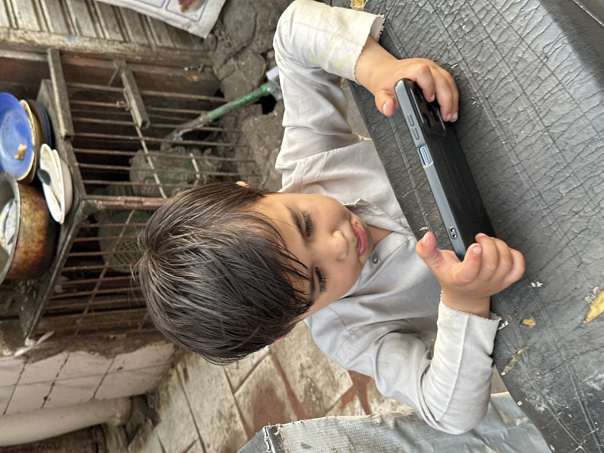 A child wearing a light-colored shirt holds a black smartphone on a dark gray surface in an urban environment. The setting includes weathered concrete, visible in the lower-left, and a dish rack with plates and pots is positioned to the left of the child. The scene appears to be outdoors in Quetta City Tehsil, Pakistan, judging by the building materials, objects present, and the light in the image.