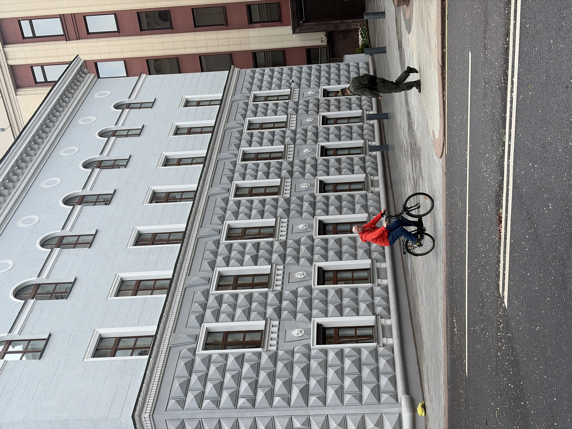 The image depicts a street scene in Minsk, Belarus. A man in a red jacket and jeans is riding a bicycle on the sidewalk. Next to him, another man in a dark green outfit is walking on the sidewalk. The street is paved with asphalt and has double white lines marking lanes. The sidewalk appears to be a mix of concrete and decorative paving stones.
The backdrop is a tall building with a facade of different styles. One side of the building has arched windows with simple white frames. The other part of the building has a textured exterior with windows and pyramid shapes. The top of the building has elements with circular decoration and molding. Another taller building with rows of rectangular windows is visible in the background.