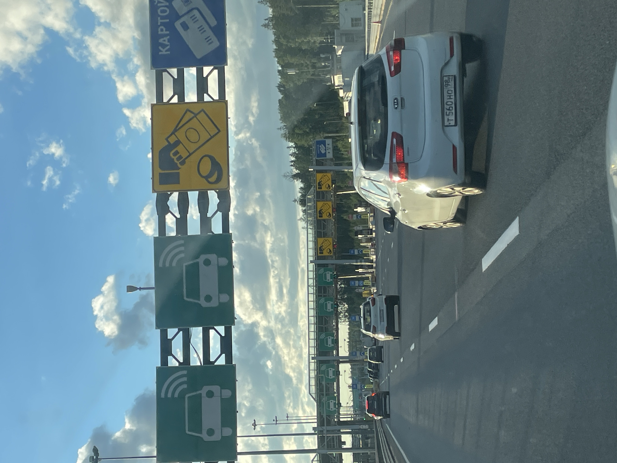 The image shows a view from inside a car driving on a highway towards a toll collection point. In the foreground, a white Kia hatchback is in the right lane. Several other cars are visible ahead, all traveling in the same direction. Overhead, a series of signs indicate toll lane options. The signs feature pictograms of cars with radio waves, suggesting electronic toll collection, and a sign with money and a credit card, indicating payment options. The sky is blue with scattered clouds, and trees are visible in the background. The location is identified as Pesochny, Russia.