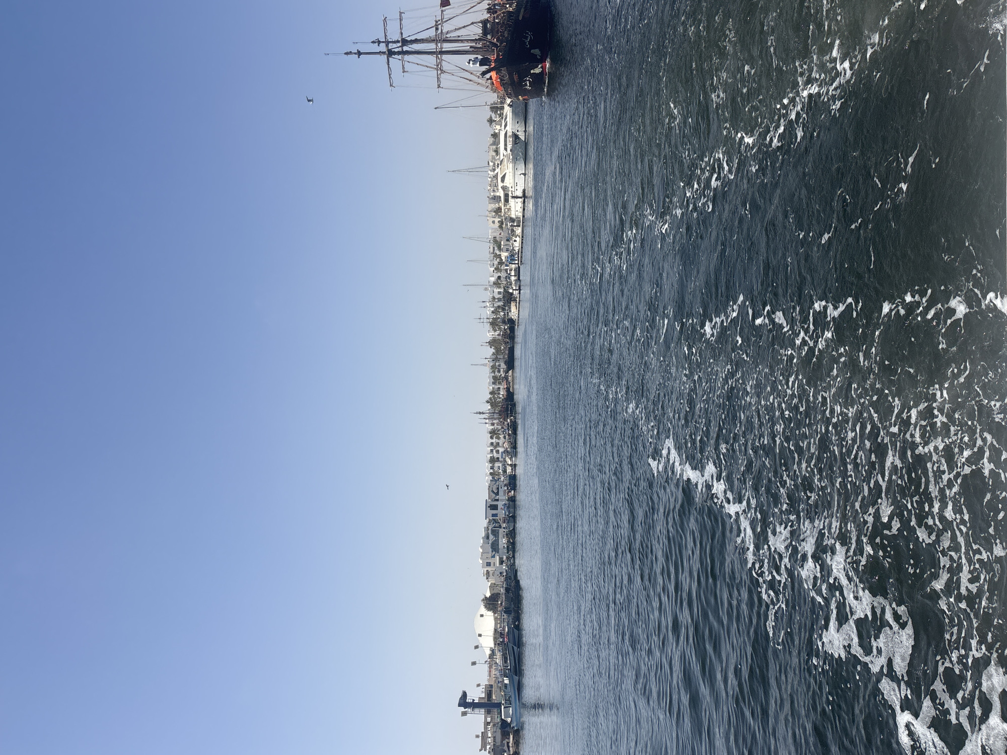 An eye-level, medium shot captures a bright, sunny day at the port of Houmt Souk, Tunisia. The scene is dominated by a clear, light blue sky above and a dark blue, choppy sea in the foreground, marked by white foam trails from a moving boat.
The port itself is lined with numerous docked boats of varying sizes, including small fishing vessels, larger yachts, and a distinctive, dark-colored, pirate-themed ship with red accents and multiple masts on the right side of the frame. The ship has Arabic writing on its hull. Several people are visible on the deck of this pirate ship.
Behind the docked boats, a strip of buildings with white facades and distinctive architecture forms the coastline. Palm trees are scattered amongst the structures. A large, white domed building is visible in the middle distance, as well as a tall, industrial-looking structure with lights near the left edge of the frame. A single bird is seen flying in the sky. The overall impression is a bustling maritime scene under a serene sky.