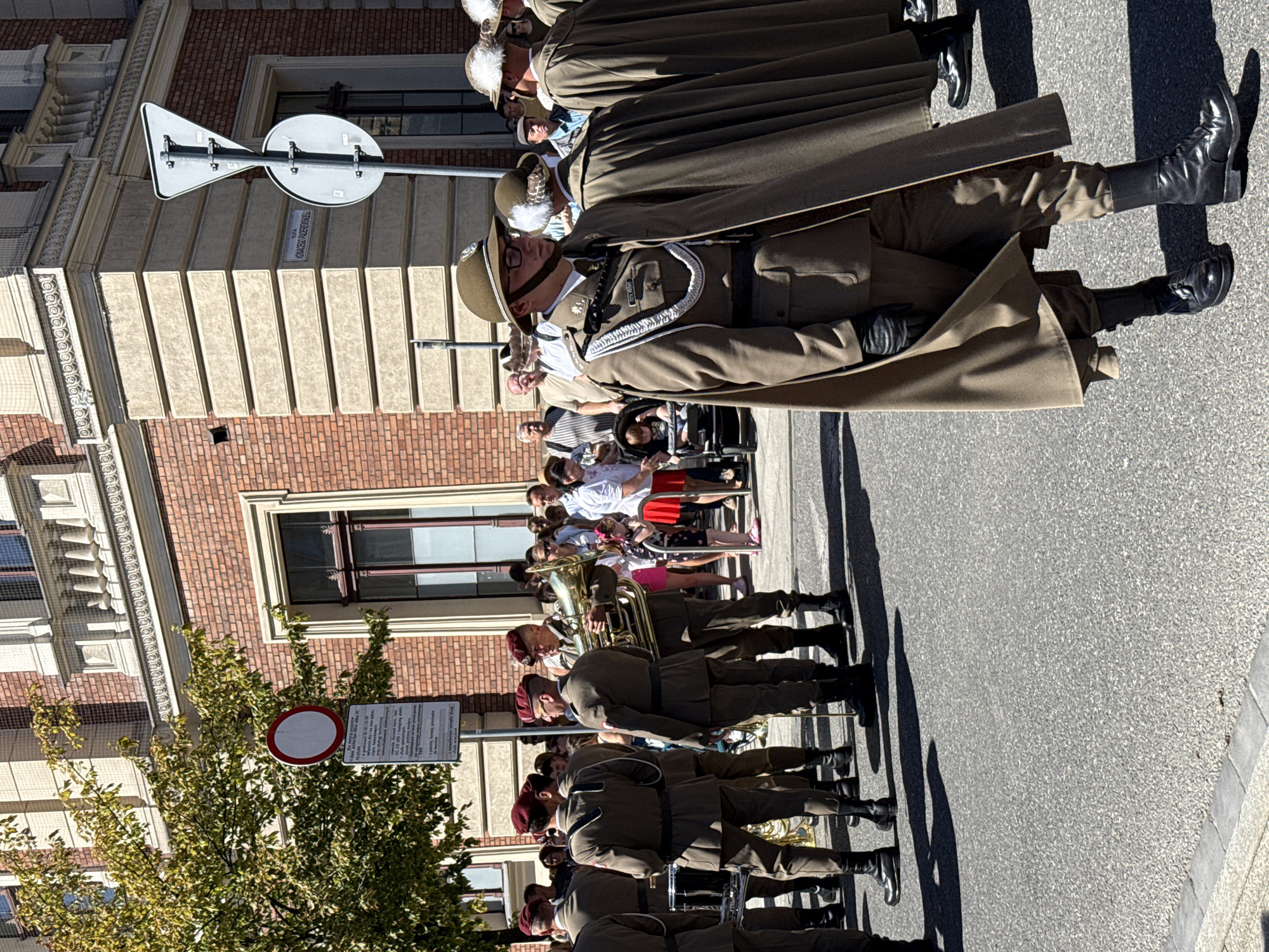 The image captures a sunny outdoor scene, likely a parade or ceremonial event, on a street in Krakow, Poland. In the foreground, a group of individuals dressed in military-style uniforms are the primary focus. They wear olive-green tunics with distinguished embellishments, dark trousers, and black boots. Several individuals are seen with long, brown cloaks draped over their shoulders. One person in the front is wearing a helmet and glasses, and another is holding a large brass instrument, possibly a tuba, suggesting a musical component to the event. Behind them, a crowd of onlookers, including men, women, and children, are gathered, some observing the uniformed group, others looking in different directions. A baby in a stroller is also visible in the crowd.
The background features a large brick building with prominent architectural details, including decorative cornices and framed windows, indicative of historical architecture. Street signs are also visible, with one clearly showing "ULICA IGNACEGO PADEREWSKIEGO," identifying the location as Ignacy Paderewski Street. Another sign, a circular red and white sign, likely a traffic regulation, is attached to a pole and partially obscured by tree branches. The ground is paved with asphalt, with a visible grey border. The overall atmosphere suggests a public event taking place on a pleasant day.
