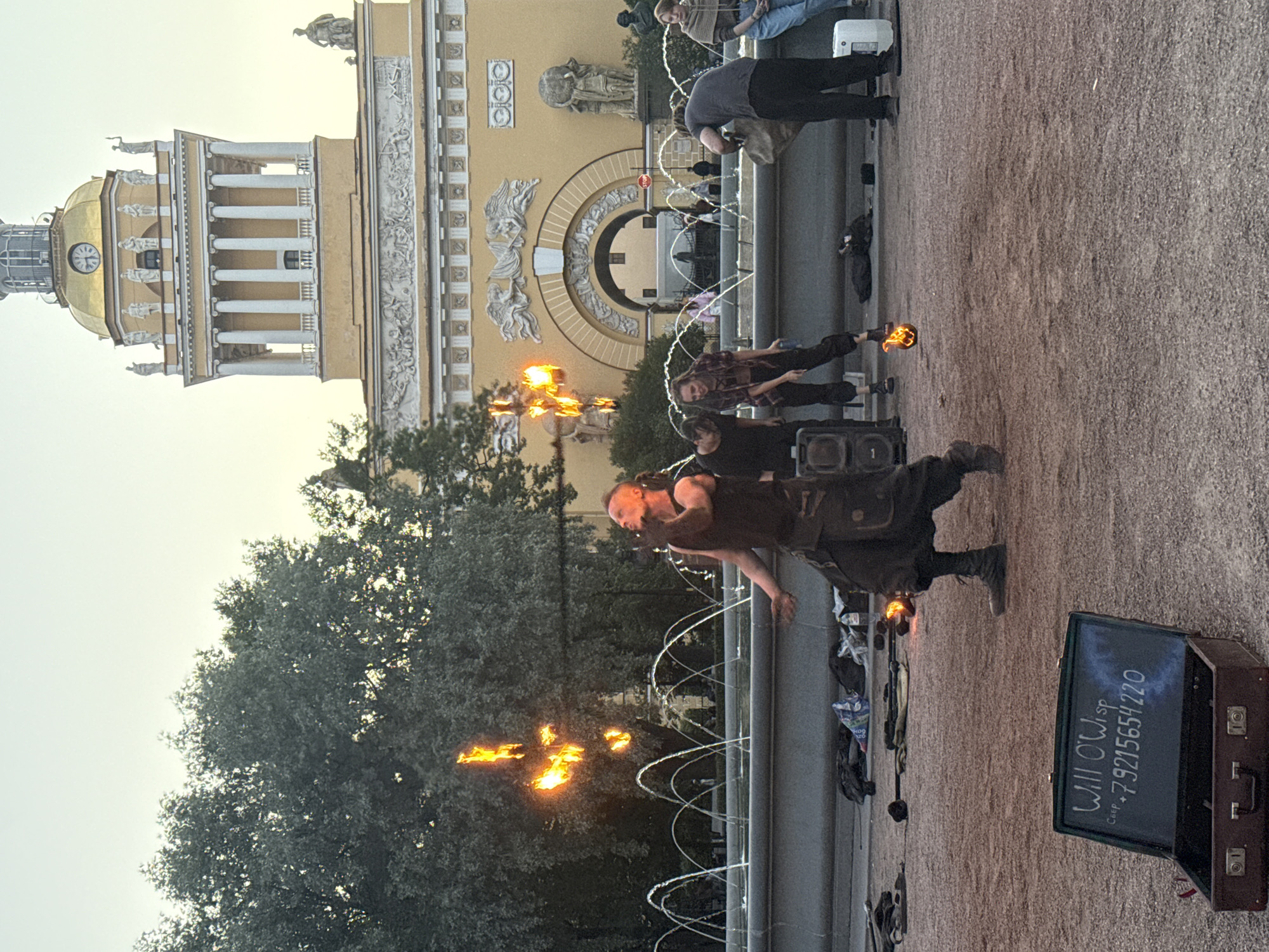 A fire performer is juggling flaming objects in front of a large fountain in Palace Square, Saint Petersburg, Russia. The performer, wearing dark, utilitarian clothing, is in the center of the frame, with flames arcing above his head. Other individuals are visible in the background, some watching the performance and others engaged in setting up or packing away equipment. Behind the fountain, a large yellow building with classical architectural features and a golden dome stands prominently. The sky is overcast, suggesting either early morning or late evening. A vintage-style suitcase with chalk writing on its lid is open in the foreground, displaying the text "Will O'Wisp" and a phone number. The ground is covered with gravel and dirt.