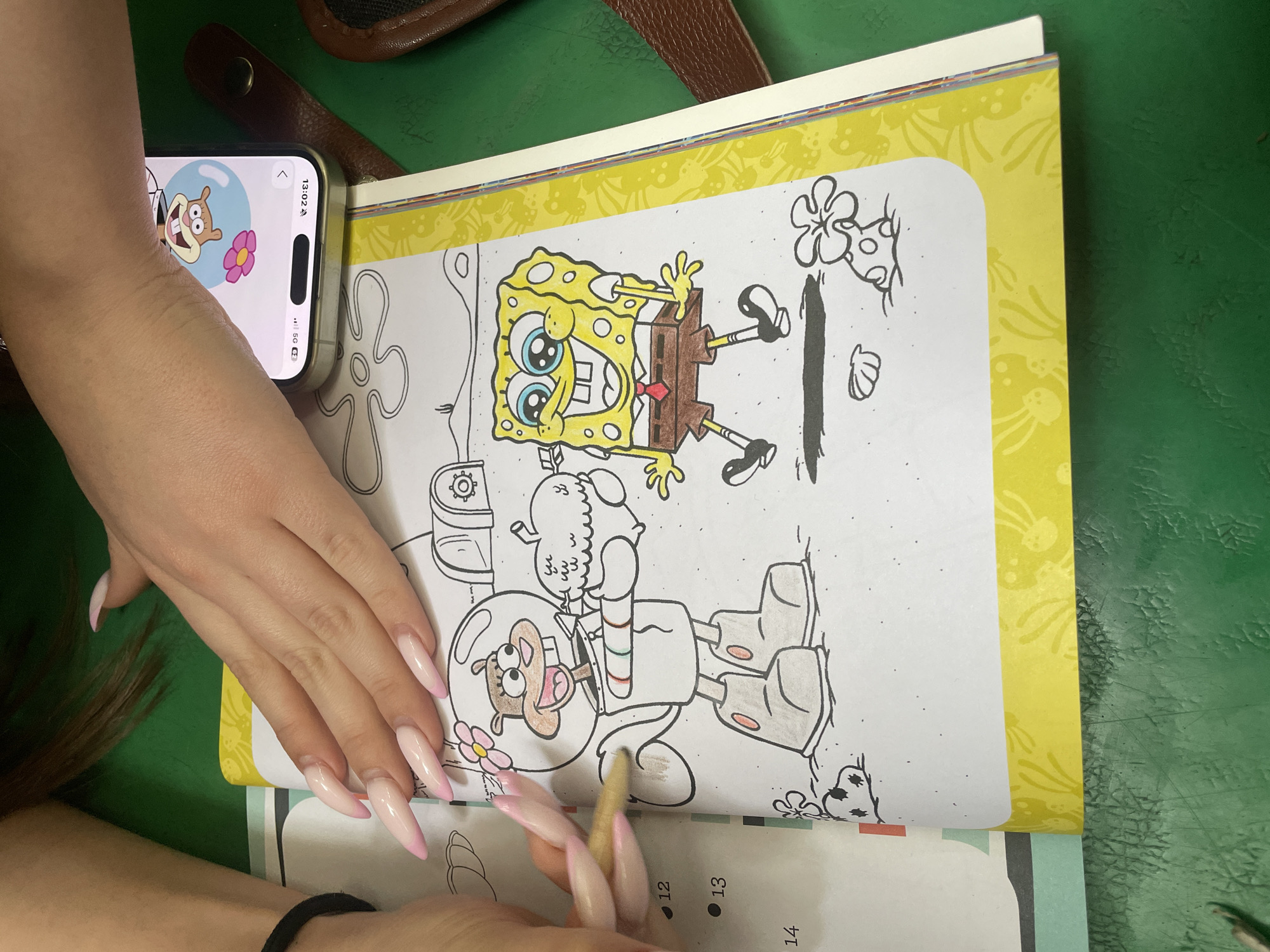 A person is coloring in a SpongeBob SquarePants coloring book. Sandy Cheeks is being colored with a yellow crayon. The coloring book has a yellow border with white jellyfish. A cell phone sits open next to the book, displaying an image of Sandy Cheeks. The scene is occurring on a green table. The location is not discernible.