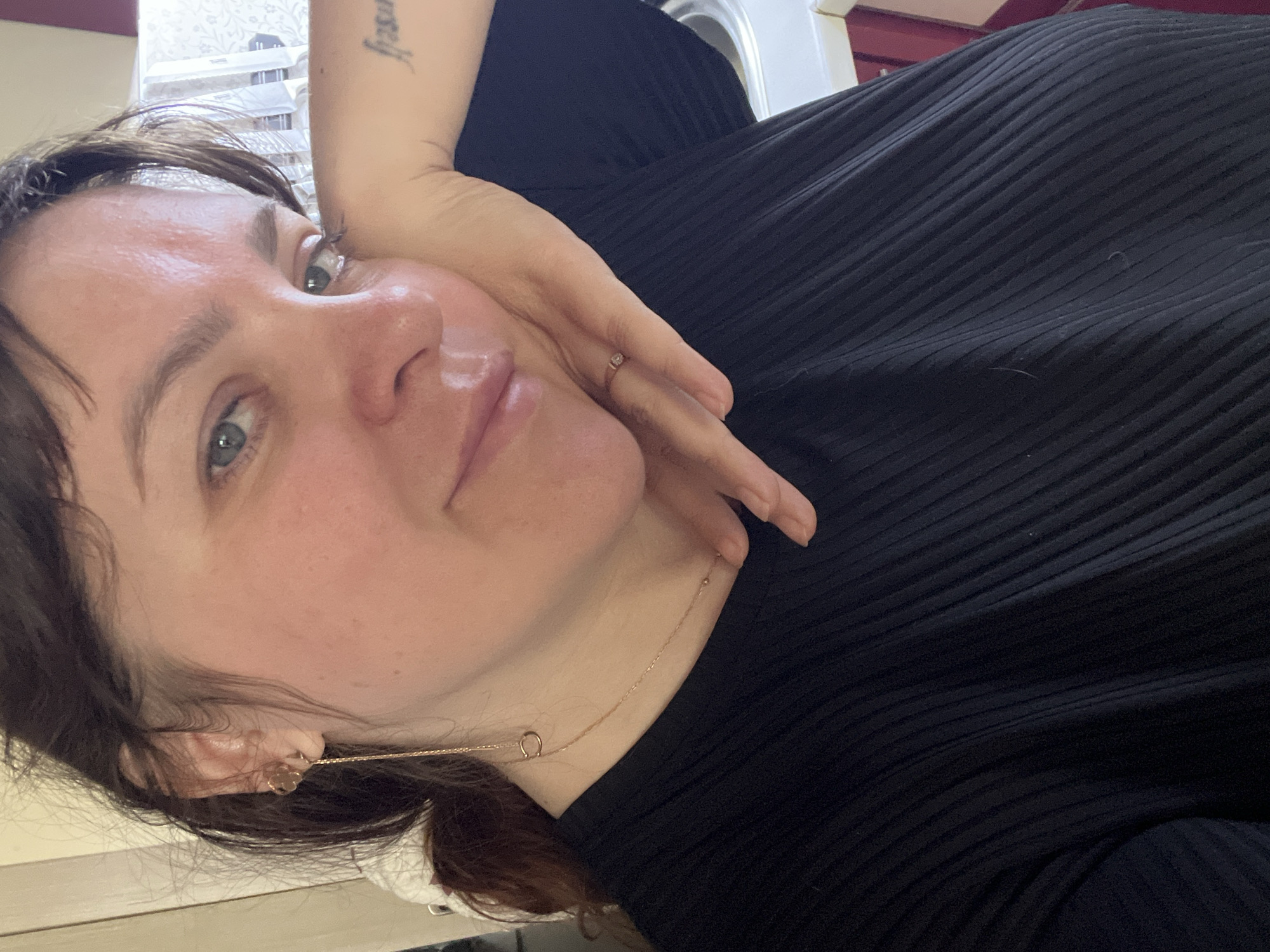 A medium shot captures a woman in close-up, resting her head on her left hand while looking directly at the camera. She has dark hair pulled back and is wearing a black, ribbed t-shirt and a delicate gold necklace. A gold earring with a chain drapes down from her left ear. A ring with a small gem adorns her right index finger, and a tattoo is visible on her right forearm. The background is blurred, but suggests an indoor setting with natural light. There is no discernible information about the location in Noviy, Russia.