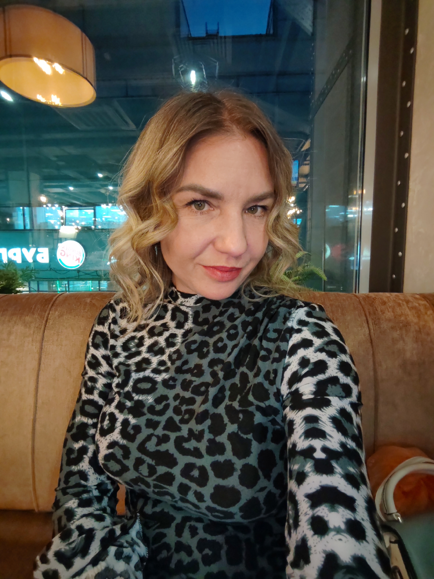 The image features a woman posing for a direct-to-camera portrait, likely a selfie, while seated indoors at night. She has shoulder-length, wavy blonde hair and a subtle smile, looking directly at the viewer with light brown or hazel eyes. She is wearing a long-sleeved, high-necked top with a distinctive black and gray leopard print pattern.
The woman is seated on a plush, brown upholstered booth or sofa. Behind her, a large window offers a view of an urban landscape at night, characterized by a dark sky and various illuminated structures and signs. In the upper left, a warm-toned, cylindrical ceiling lamp casts a soft glow.
Notable details include the time of day, which is evening or night, indicated by the dark outdoor scene and artificial lighting both inside and outside. Through the window, reflections show parts of illuminated signs. Specifically, a white circular sign with the text "KING Burger" is partially visible, suggesting a Burger King restaurant nearby. Also visible is reversed text "ЯVA," possibly from another sign. The woman appears relaxed and composed. Given the location context, this scene is set in Saint Petersburg, Russia.