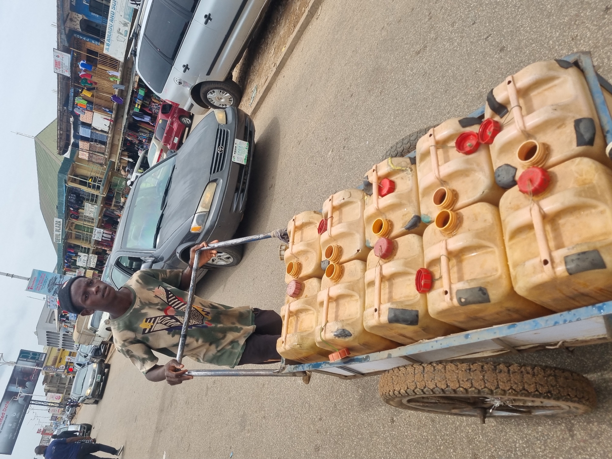 The image shows a young man pulling a two-wheeled cart laden with yellow jerrycans, likely filled with water, on a street in Jos, Nigeria. He wears a patterned t-shirt and a small black cap. The street is flanked by buildings, with visible storefronts and signs in the background, one reading "SEVIL PLAZA", another reading "A.B AMADU AND SUP." A gray Toyota Camry and other vehicles are parked along the street. The cart's tires are covered in mud. The overcast sky suggests it is daytime.