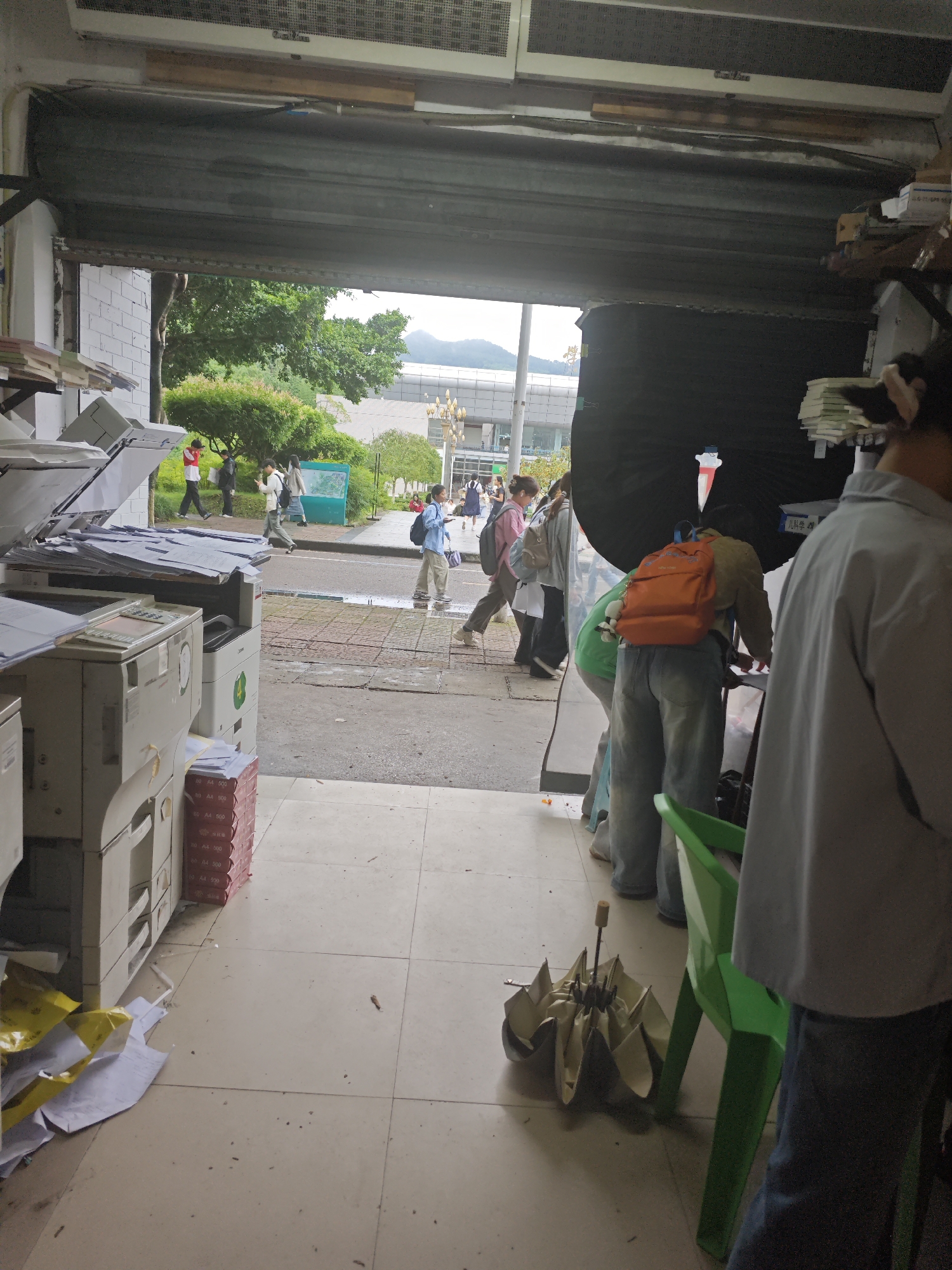 The image depicts the interior and entrance of what appears to be a print shop or copy center located in the Chongqing High-tech Zone, China. Through the open metal gate, a street scene is visible with trees, buildings, and people walking. Inside the shop, there are multiple copy machines, stacks of paper, and some discarded papers on the floor. In the foreground, a person in a light-colored shirt and dark pants stands partially in view, and a green plastic chair is positioned to their right. On the floor next to the chair lies a folded umbrella. Further into the shop, another person is seen wearing a green shirt, blue jeans, and an orange backpack, leaning over a surface. Several other individuals are visible in the street outside, some carrying backpacks, suggesting they might be students or commuters. The overall environment appears somewhat cluttered, with a mix of business equipment and everyday items.