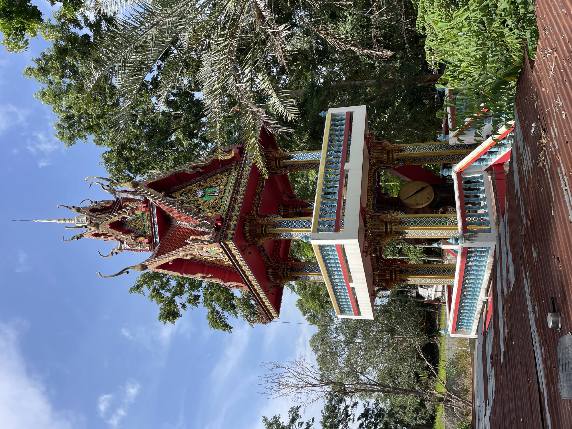 An ornate, multi-tiered bell tower stands prominently in a tropical outdoor setting under a bright blue sky with scattered clouds. The tower is painted in vibrant colors, featuring red, gold, and blue accents. It has a pagoda-like roof with intricate carvings and decorative elements. A balcony with a white railing, accented with blue and red spindles, encircles the second tier. Visible through the open sides of the tower on the second level is a large, circular drum, likely a gong. The tower is surrounded by lush green trees and foliage, including palm fronds. In the foreground, a rusty, corrugated metal roof covers a structure, and a small metal pot is visible on its surface. The scene suggests a temple or a place of worship, possibly located in Pattaya City, Thailand, given the architectural style and the tropical vegetation. There are no people visible in the image.