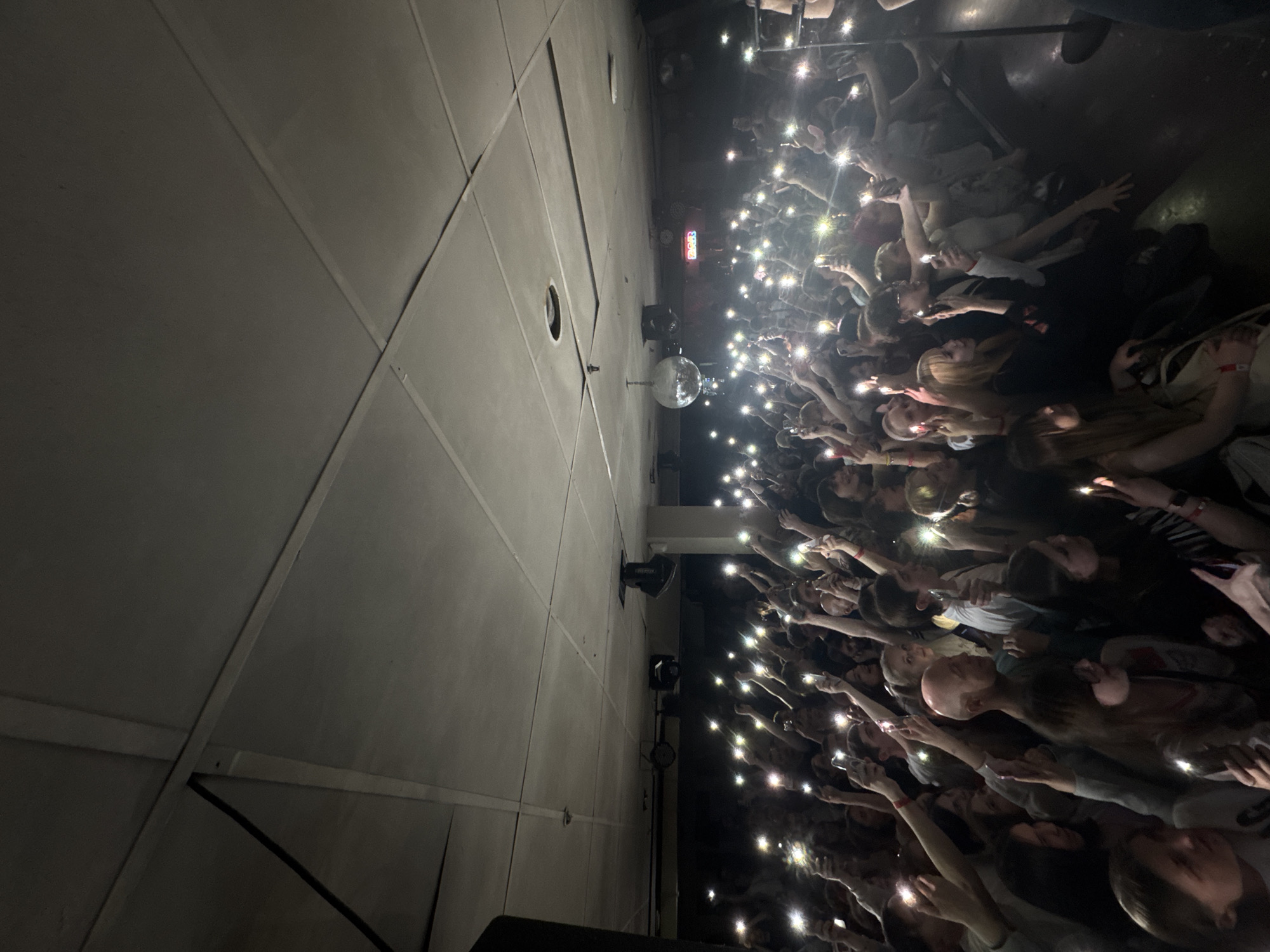 This image captures a vibrant and densely packed indoor event, likely a concert or performance. The scene is dominated by a large crowd of people, many of whom have their arms raised, holding cell phones with their flashlights activated. These numerous points of light create a starry effect across the dark venue, illuminating parts of the attendees' faces and hair.
The photograph is taken from an elevated perspective, looking down and across the space. To the left, a large, light-colored ceiling or wall structure with visible panels and seams occupies about half of the frame. Two circular cutouts are visible in this structure, possibly for speakers or vents. A small disco ball hangs near the center of the image, reflecting some light. Below the ceiling structure, various stage lights or equipment are mounted.
The activity clearly depicts an engaged audience at a live event, using their phone flashlights in a common gesture of appreciation or participation during a performance. The atmosphere appears dark, indicative of nighttime or an indoor venue with controlled lighting, where the cell phone lights provide significant illumination.
A notable detail is a glowing red/pink neon sign visible in the background near the ceiling, spelling out "RAGER". No specific weather conditions are discernible, and no direct cues point to Krasnoyarsk within the image itself. The overall emotion conveyed is one of engagement and excitement.