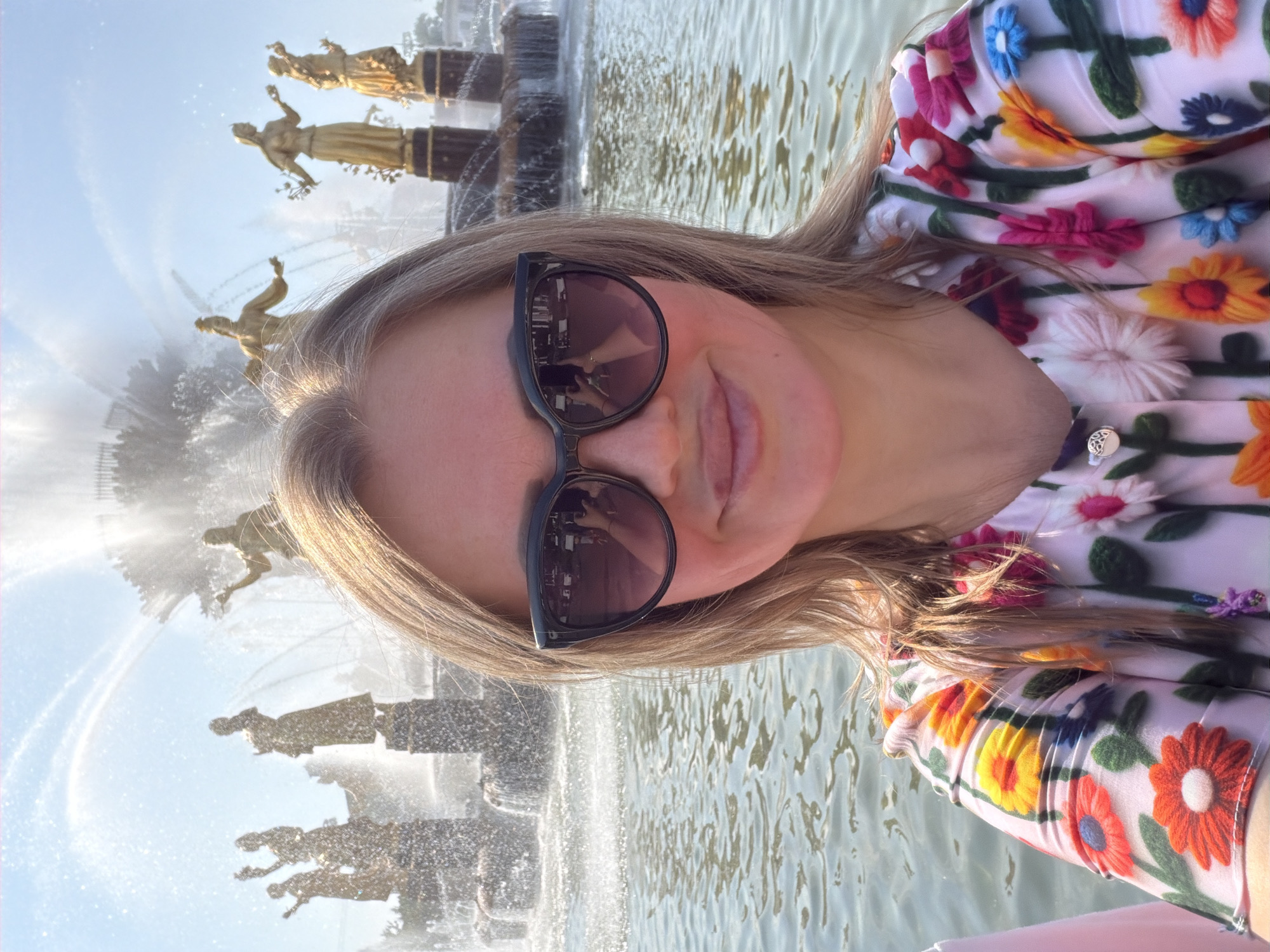 A woman with blonde hair and sunglasses takes a selfie in front of the Friendship of Peoples Fountain at VDNKh in Moscow, Russia. The fountain features several golden statues of women holding various objects, surrounded by jets of water and illuminated by sunlight. The woman is wearing a colorful floral print shirt with 3D flower embellishments. The water in the fountain is rippling, and the sky is clear and blue.