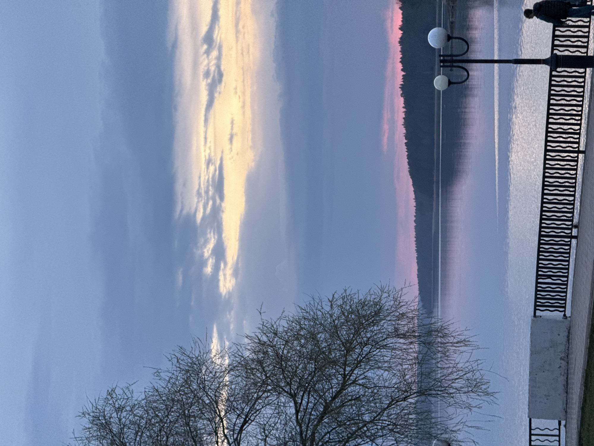 The image depicts a waterfront scene in Votkinsk, Russia, likely captured during twilight. A body of water reflects the sky, which is streaked with both dark and light-colored clouds. A strip of land with trees can be seen across the water. A black metal railing runs along a concrete walkway. A lamppost with two spherical lamps is positioned near the railing. A single person is visible in the top right corner of the frame, standing near the railing. Leafless trees are silhouetted in the foreground.