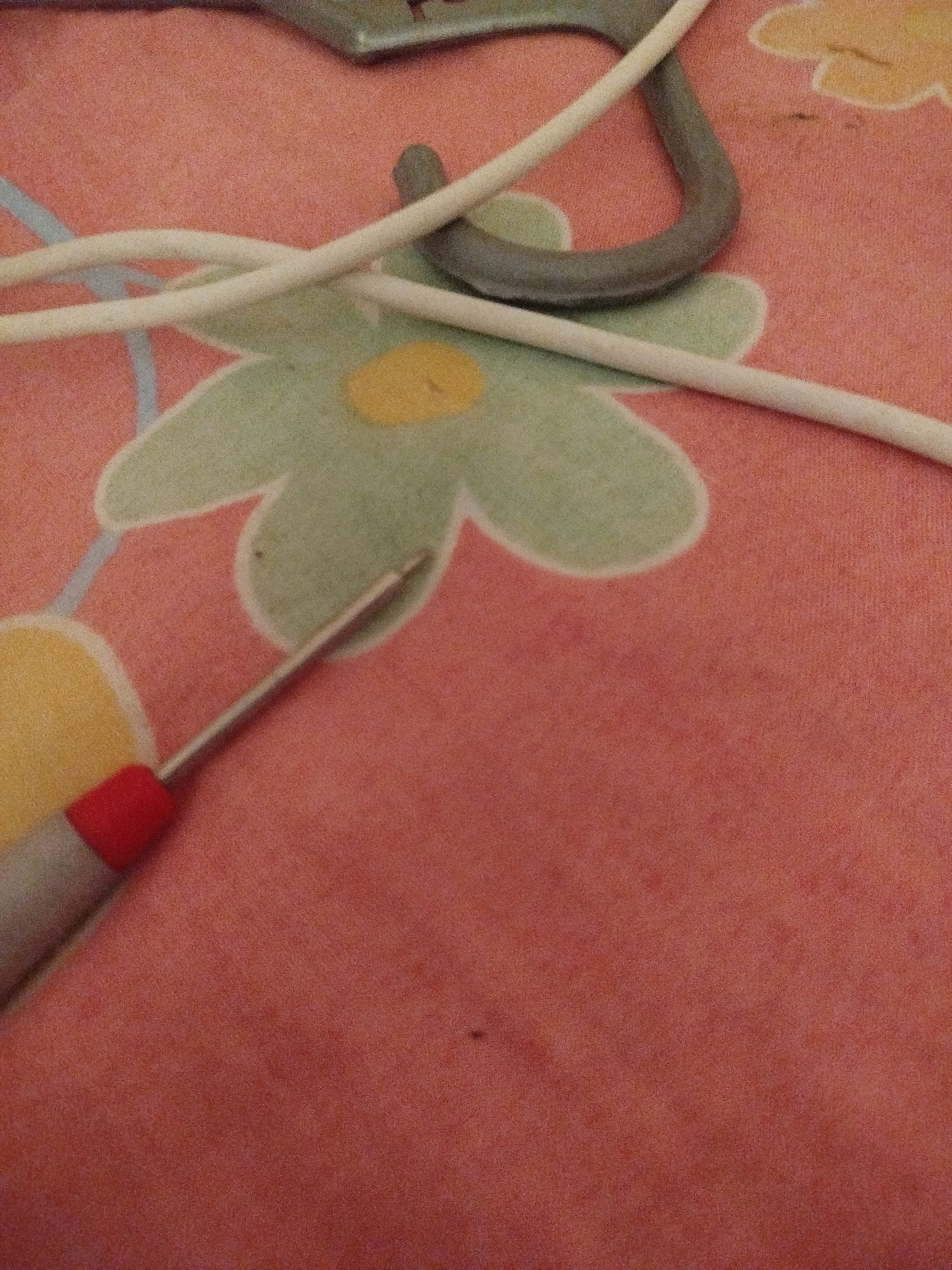 The image shows a close-up view of a pink, patterned surface, possibly a tablecloth or bedspread, with a floral design in green and yellow. Several objects are present: a gray, curved metallic object, two white cables or wires, and a small screwdriver with a red handle and a metallic tip. The screwdriver is positioned diagonally, with its tip pointing towards the upper right of the frame. The background is softly focused, emphasizing the texture and pattern of the surface and the detail of the objects. There are no discernible clues within the image to indicate a specific location in Lassan, Pakistan.